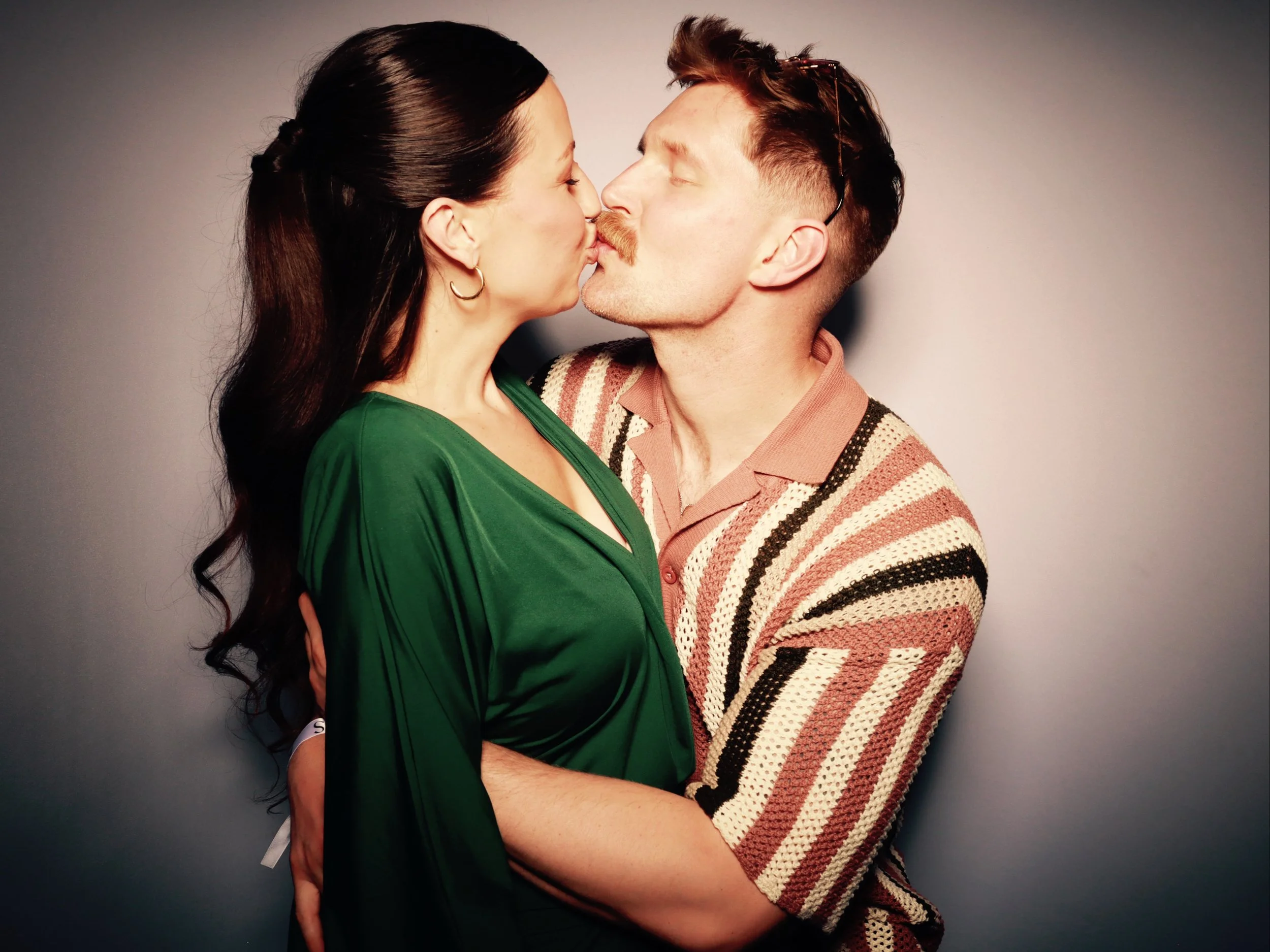 A man and woman kissing, with the woman in a green dress and the man in a patterned sweater, against a neutral background.