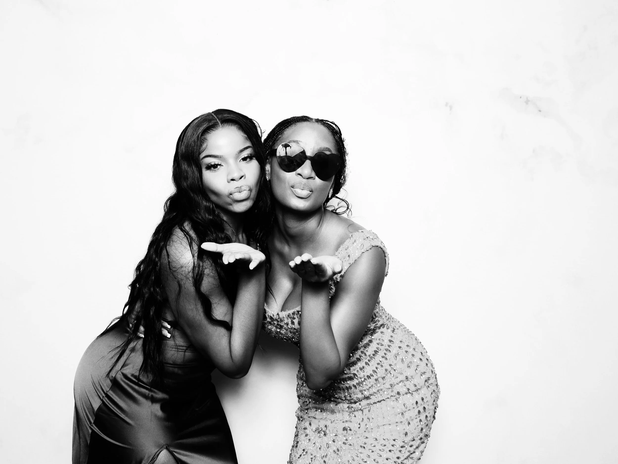 Two women blowing kisses at the camera, one wearing sunglasses and a beaded dress, the other with long wavy hair and a satin dress, against a plain white background.