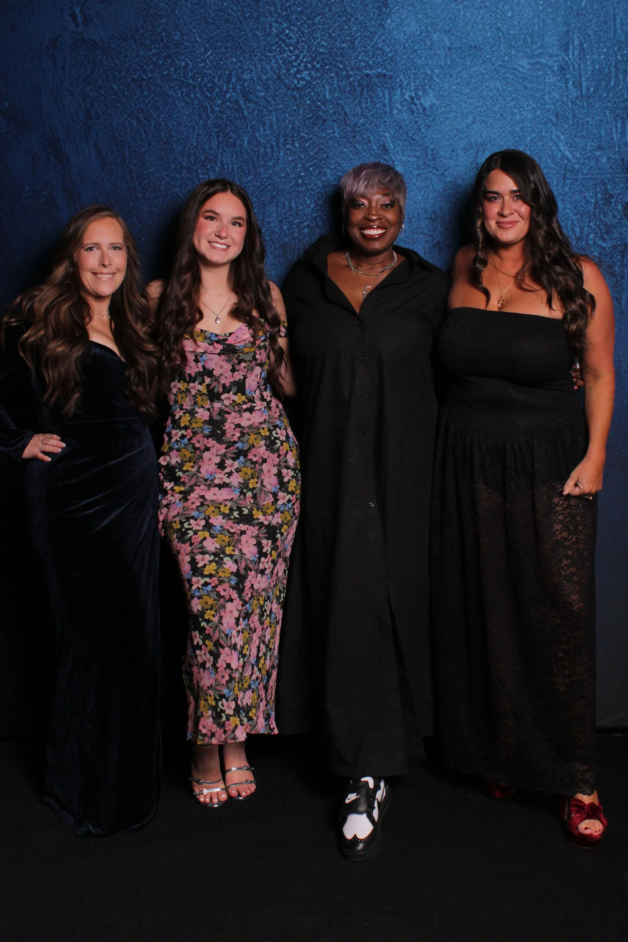 Four women standing together in front of a dark blue textured wall, dressed in formal attire for a celebration or event.