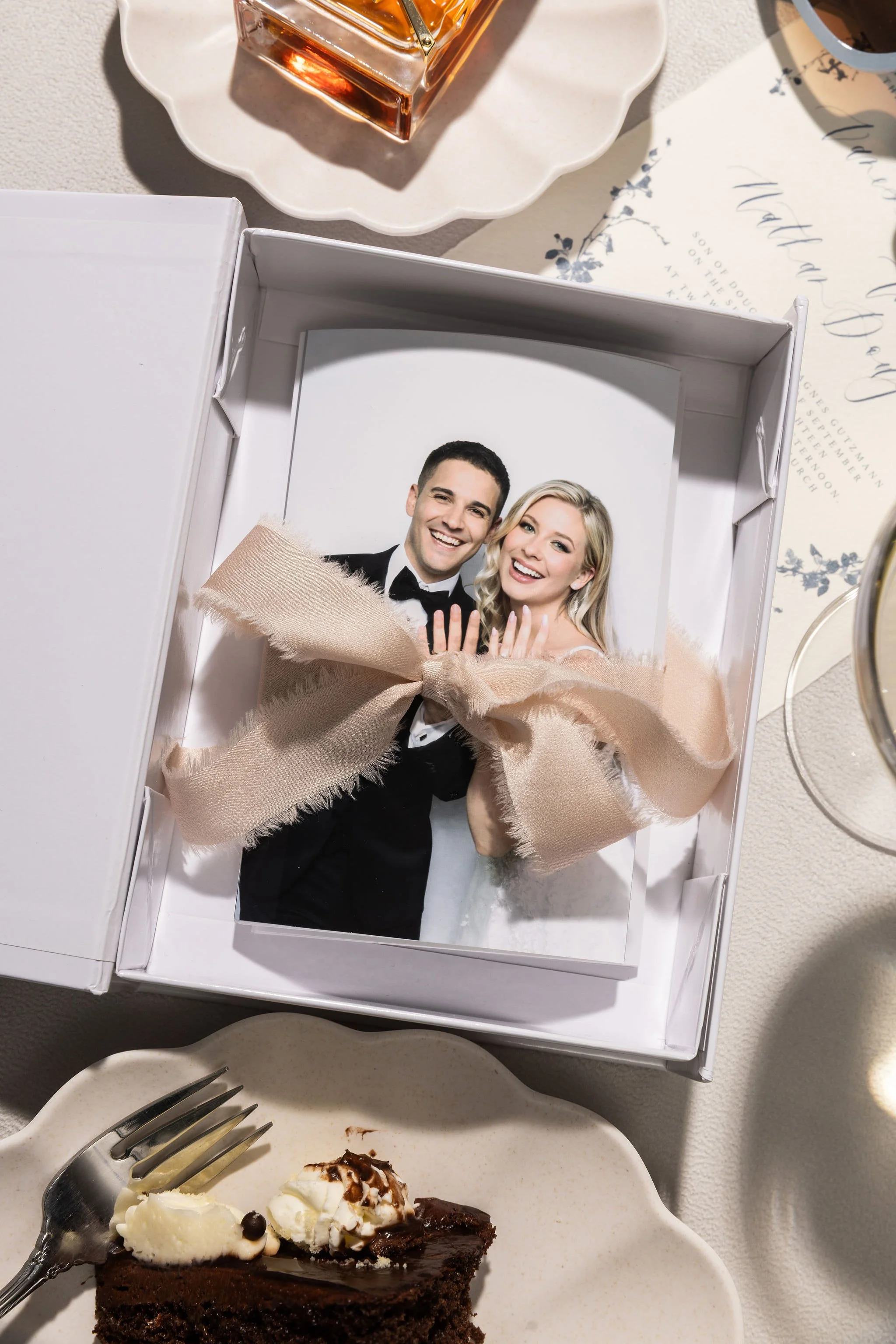 A wedding photo of a smiling couple in a box with a beige ribbon, surrounded by dinnerware and a slice of chocolate cake with whipped cream.
