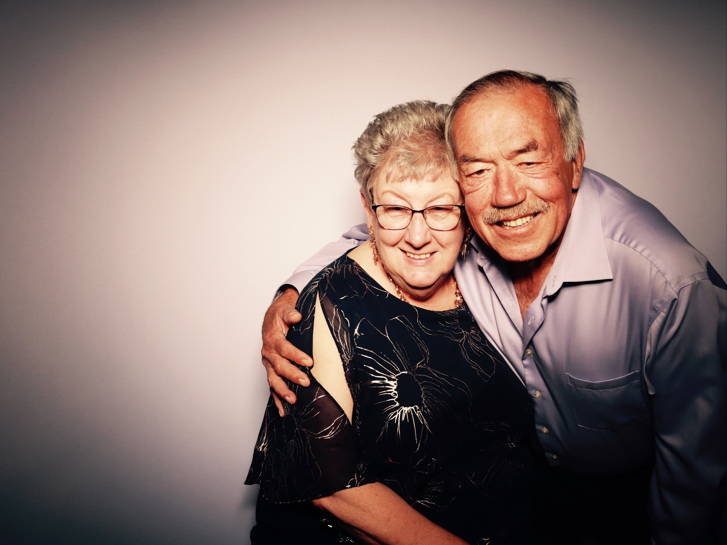 An elderly couple smiling and hugging each other against a plain background. The woman has short gray hair, glasses, and is wearing a dark dress with a floral pattern and jewelry. The man has gray hair, a mustache, and is wearing a light-colored dres