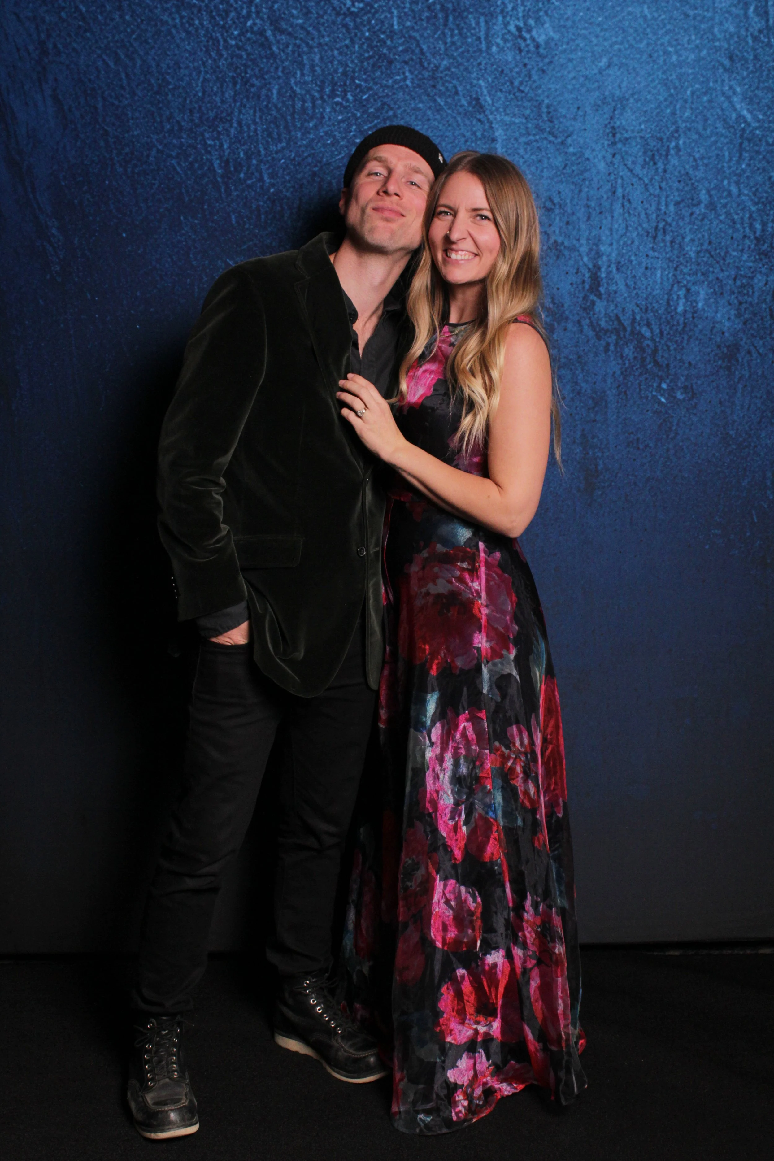 A man and woman stand close together against a blue textured background, smiling and posing for the camera.