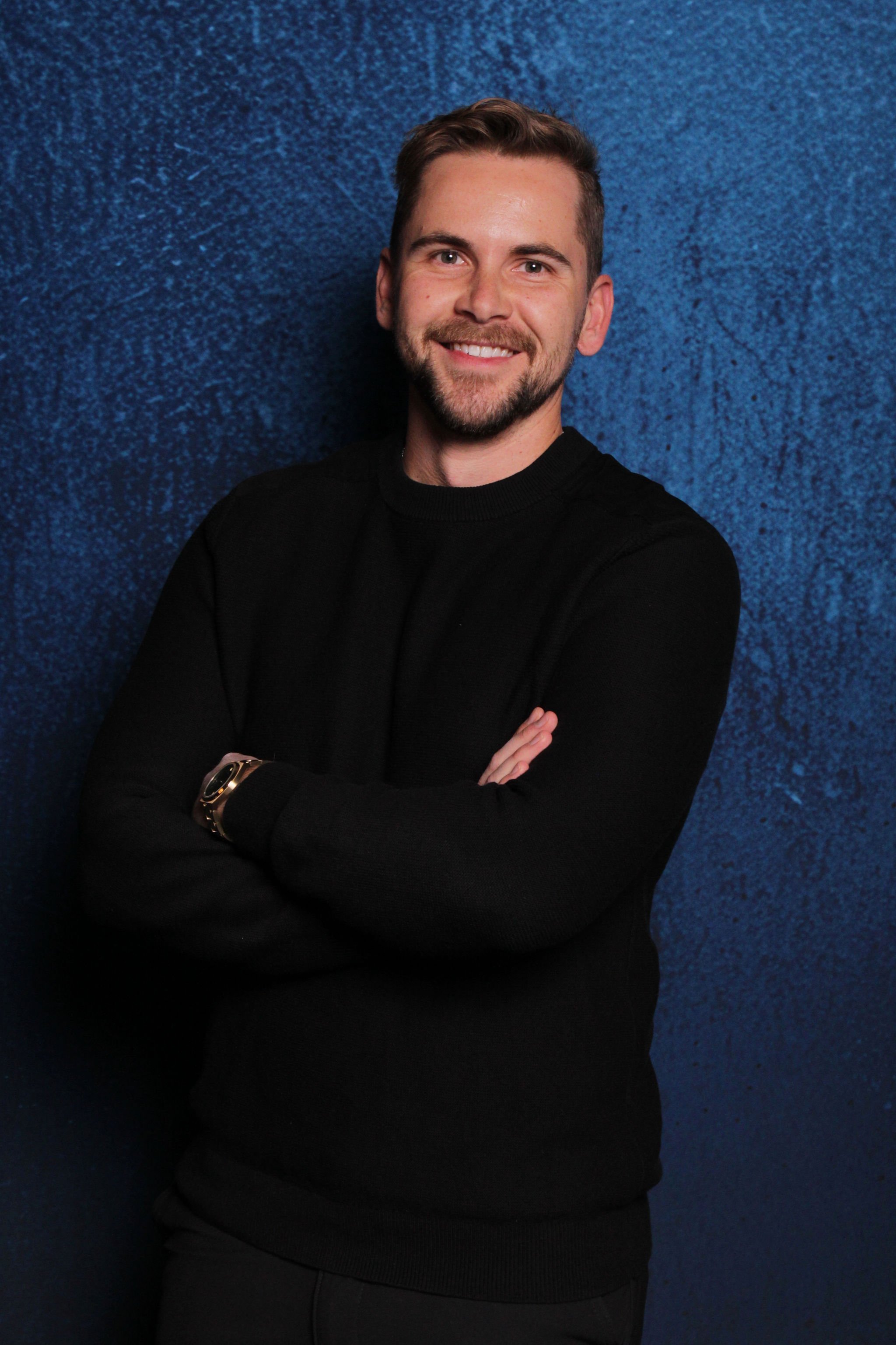 A young man with short brown hair and a beard smiling with arms crossed, wearing a black sweater, standing against a textured blue wall.