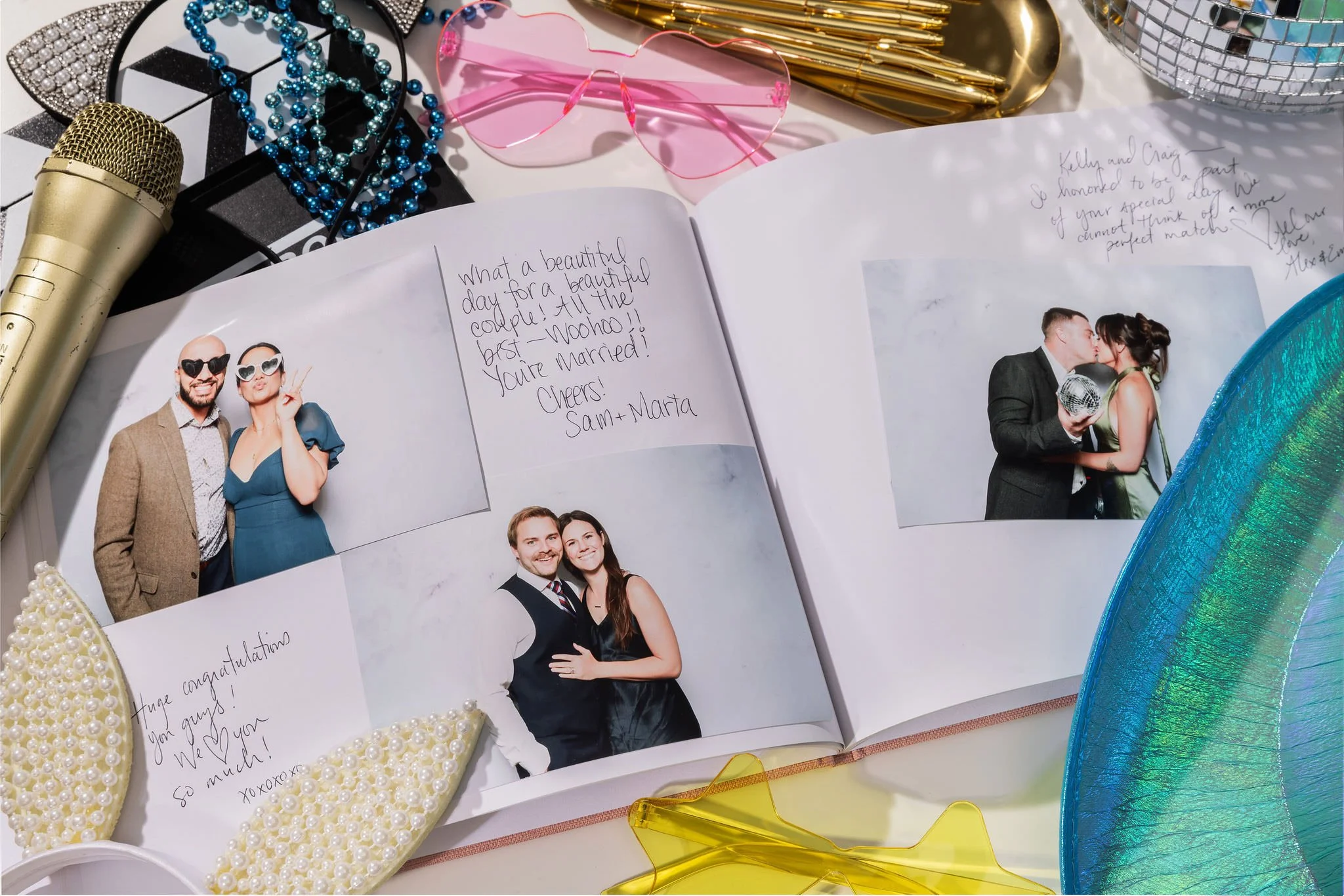 A scrapbook open to reveal wedding photos and handwritten messages, surrounded by party accessories including a gold microphone, pearl and blue beads, pink sunglasses, gold pens, a blue and green party hat, and a yellow star-shaped item.