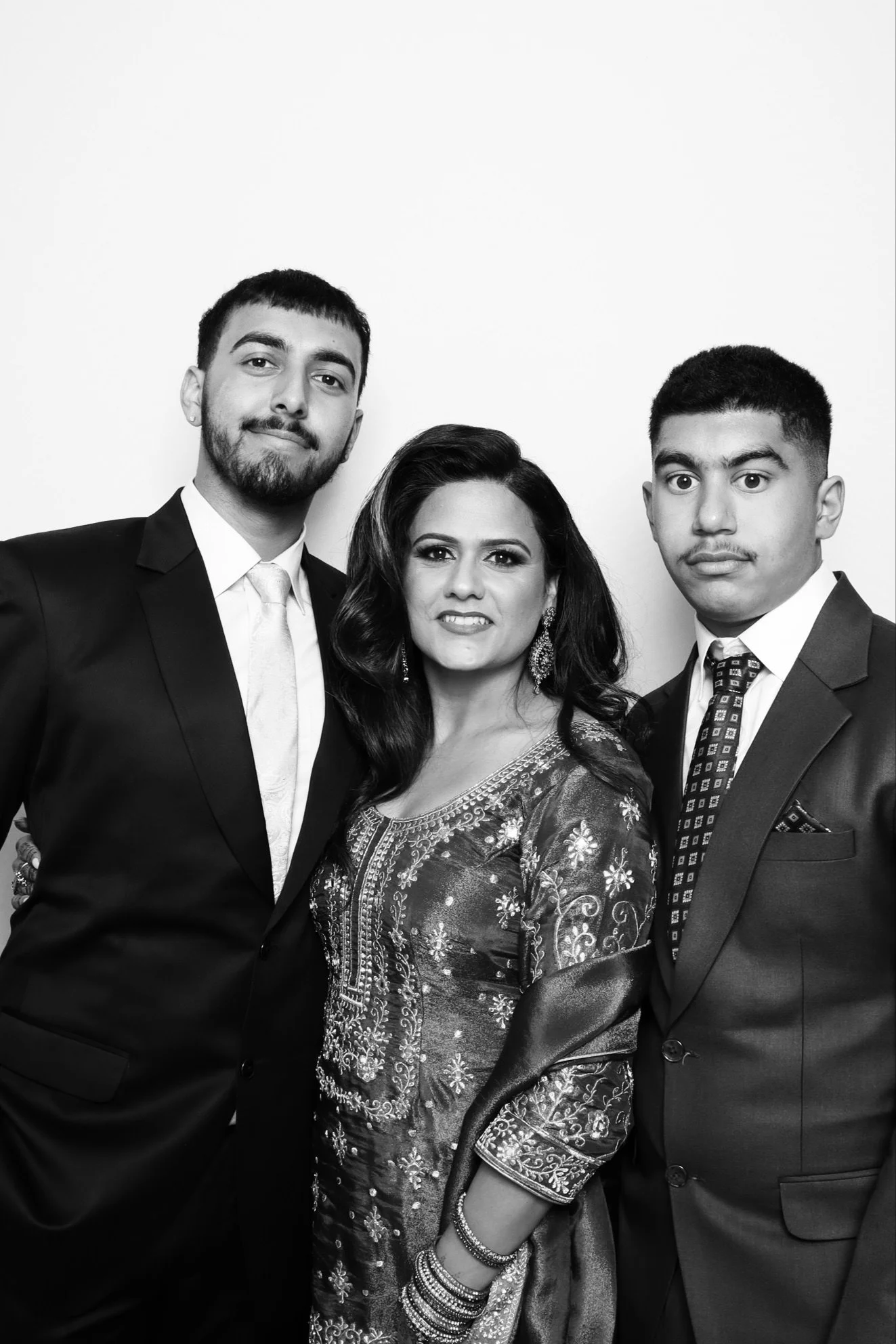 Group portrait of three individuals dressed in formal attire, posing against a plain background. The woman in the middle is wearing traditional South Asian clothing and jewelry.