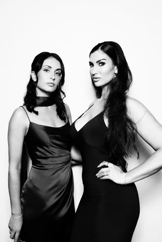 Two women in elegant black dresses standing close, facing the camera against a plain light background, one with a confident expression and the other with a neutral look.