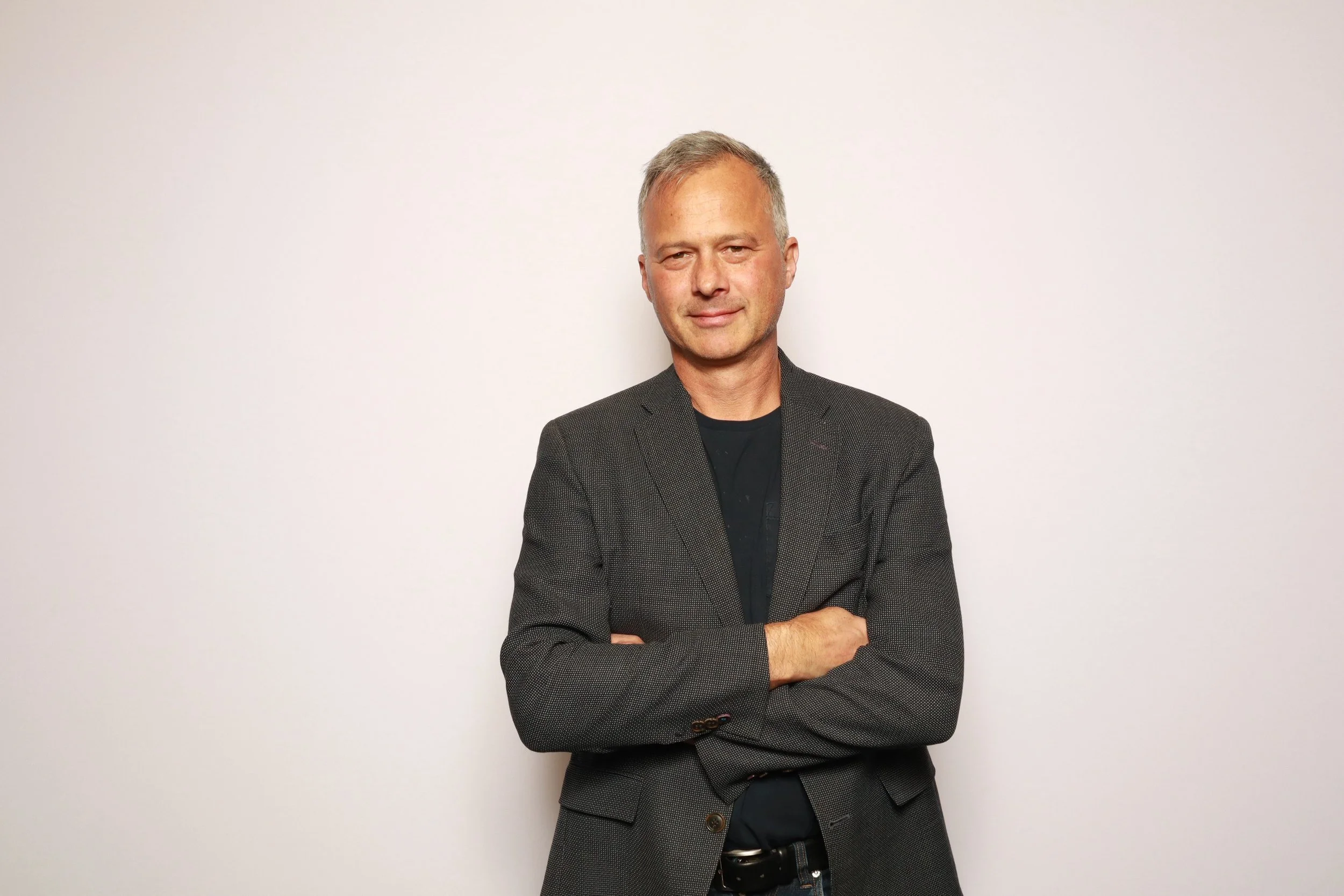 A man with blond hair and a slight smile, wearing a dark blazer and a black shirt, standing against a plain white background with arms crossed.