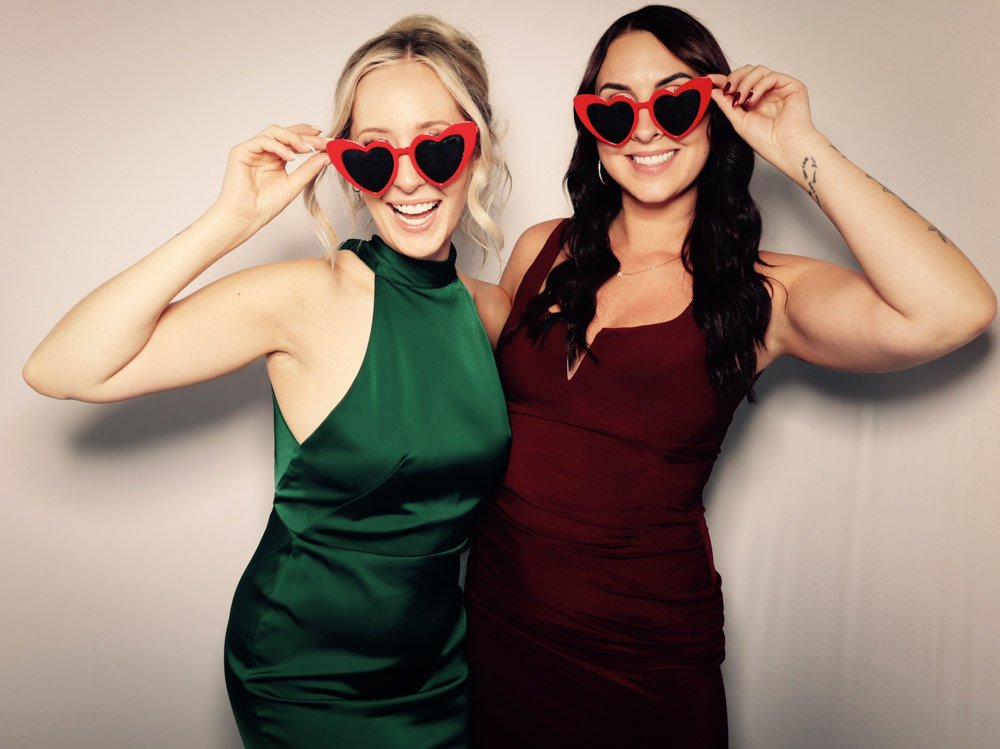 Two women smiling and wearing heart-shaped sunglasses, standing close together against a plain wall.