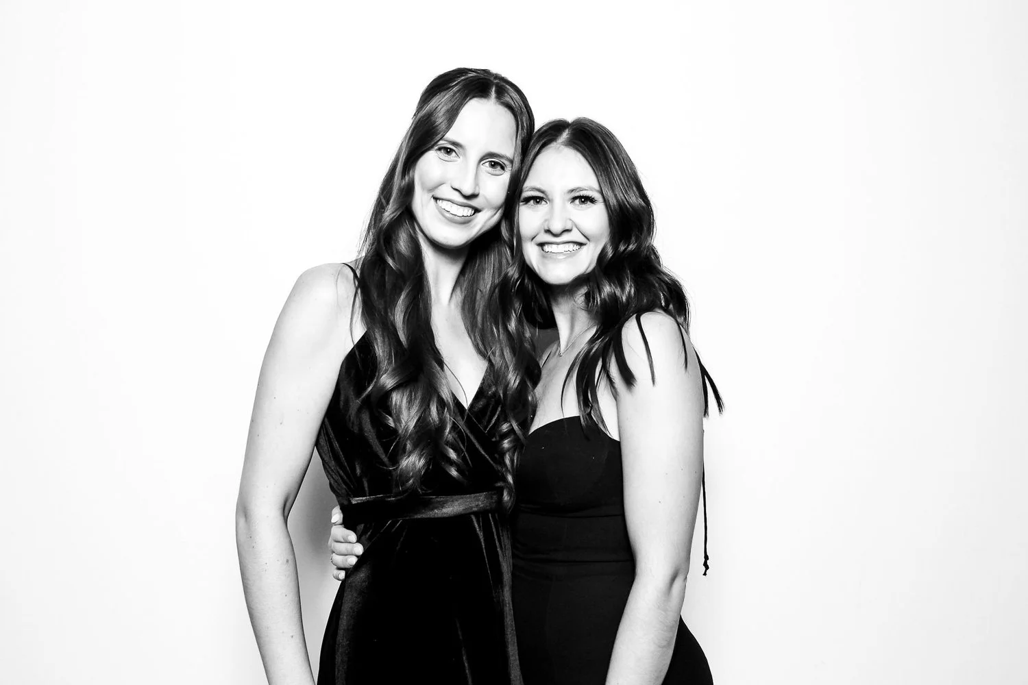 Two young women with long wavy hair smiling, standing close together against a plain background, wearing dark sleeveless dresses.