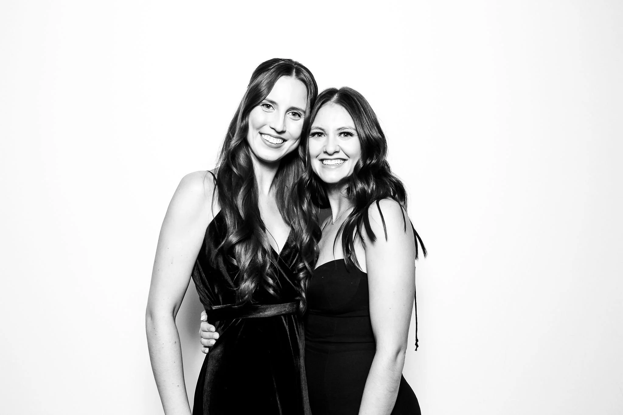 Two women with long wavy hair smiling, wearing sleeveless black dresses, standing close together against a plain light background.