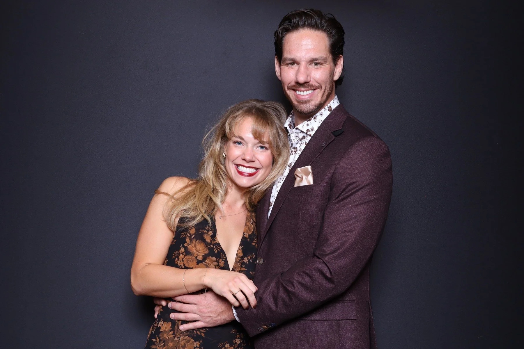 A smiling couple, a woman with blonde, wavy hair wearing a floral dress, and a man with dark hair and a beard in a suit, standing close together against a dark background.