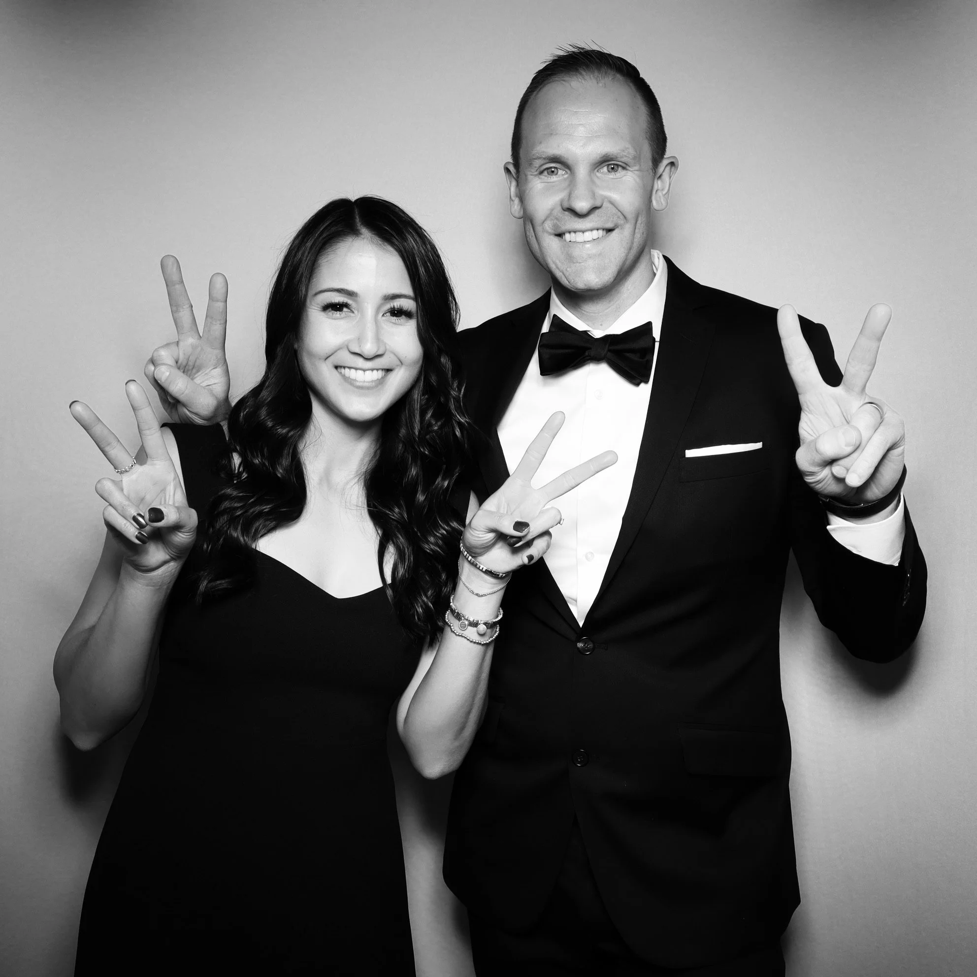 Black and white photo of a smiling man and woman in formal attire making peace signs with their hands.