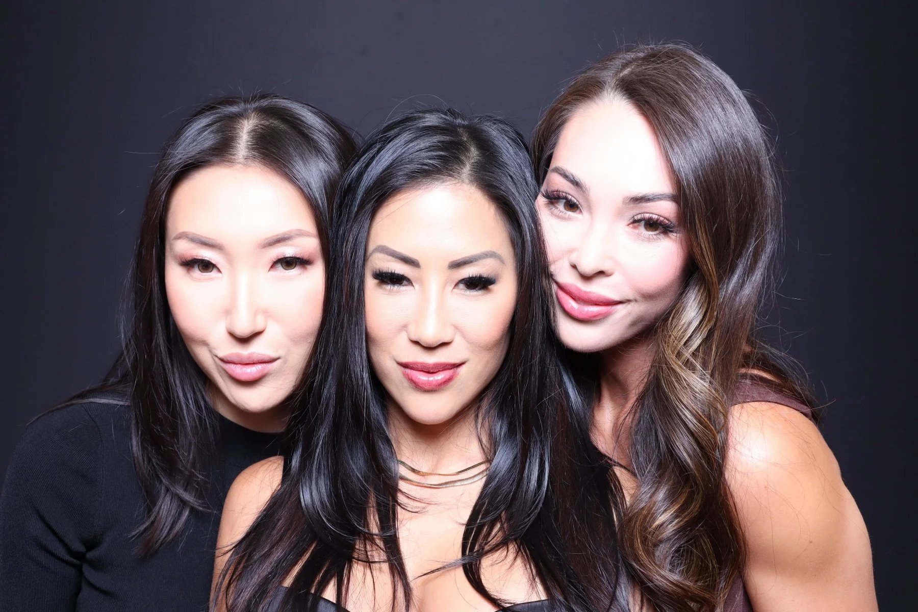 Three women with long hair, close together, smiling against a dark background.