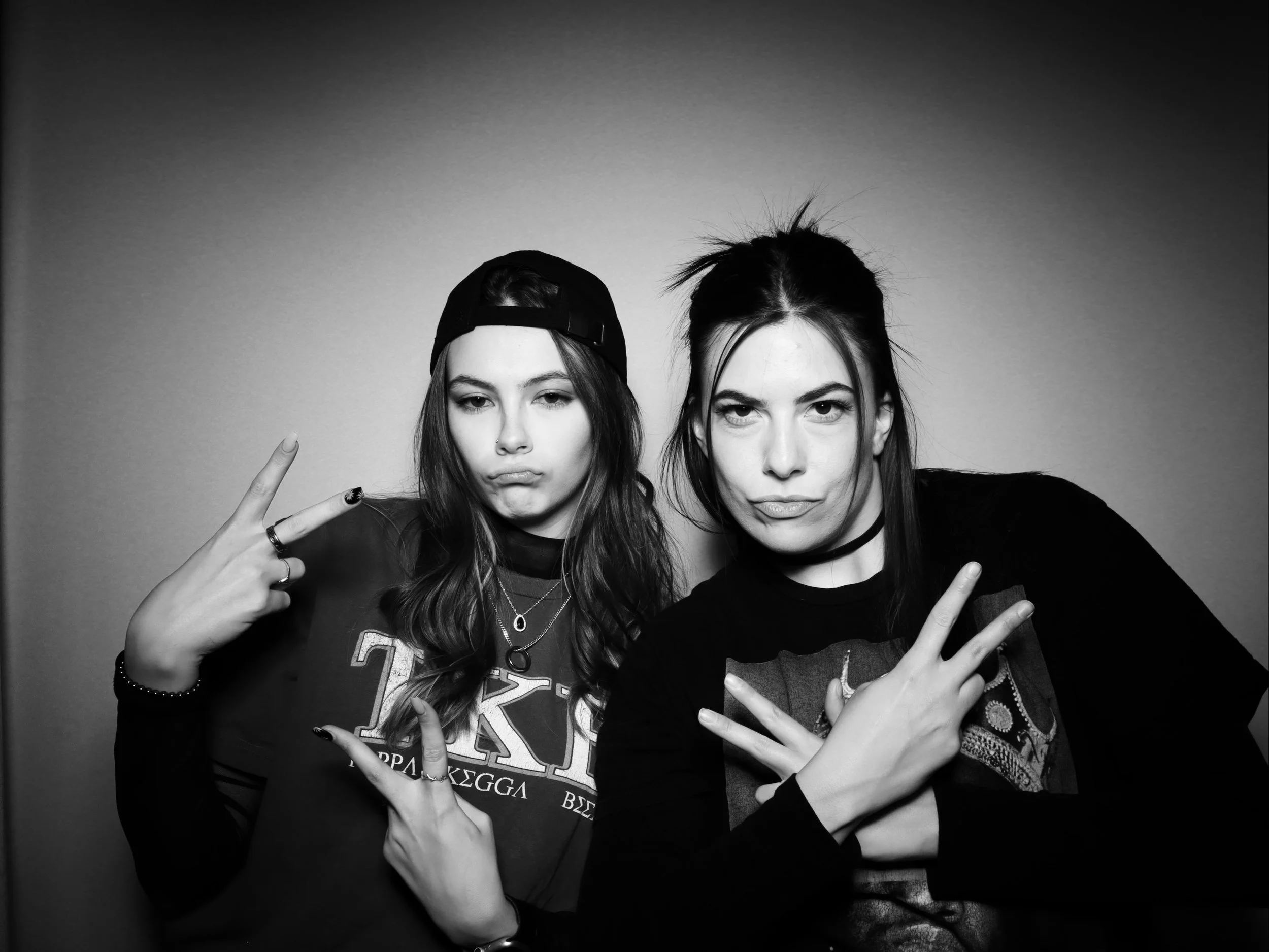 Two young women pose with defiant expressions and peace signs, one wearing a backward cap and layered necklaces, the other with a choker and styled hair, against a plain background.