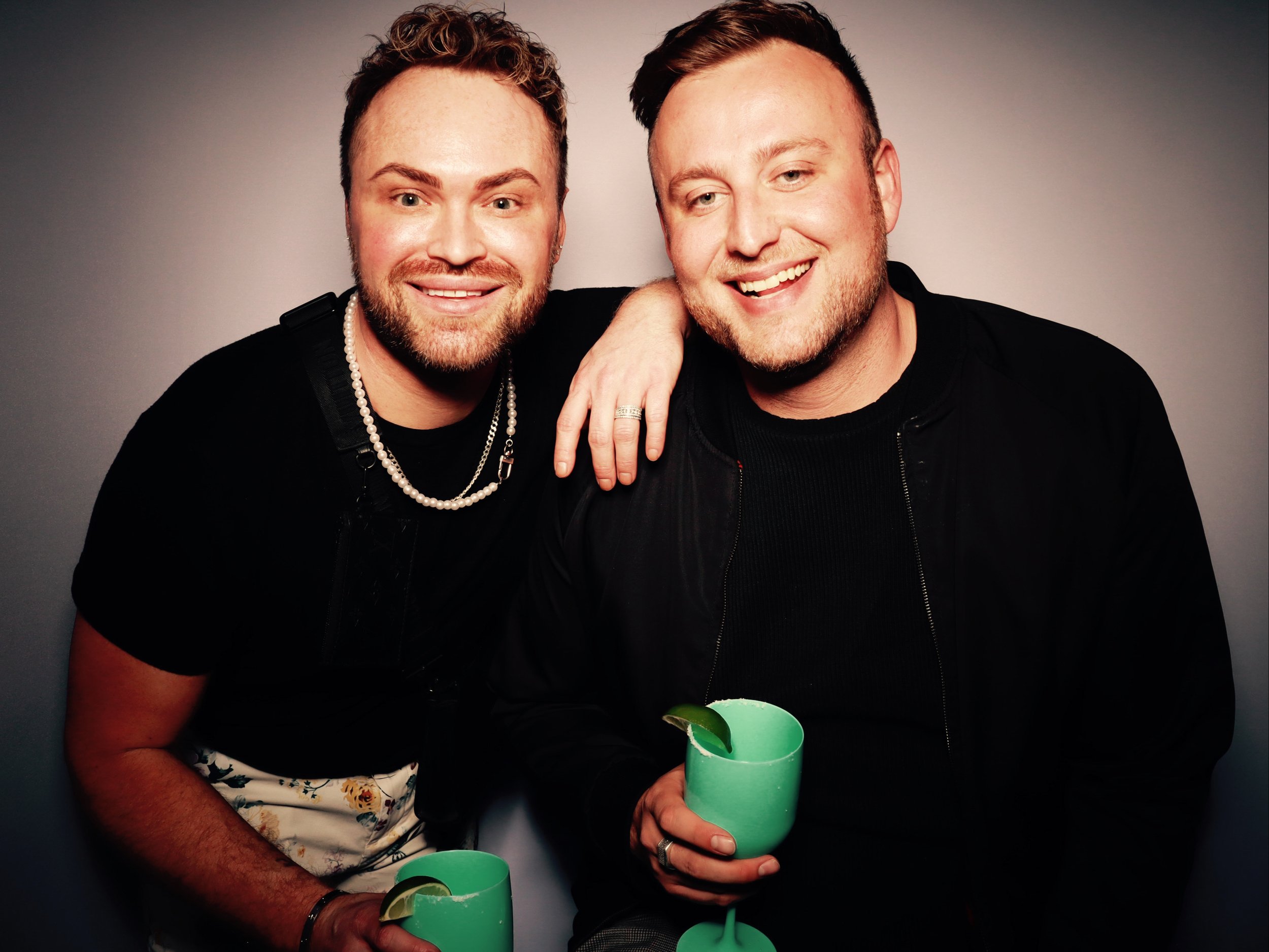 Two smiling men holding green drinks with lime slices, one has their arm around the other in a friendly pose, against a plain background.
