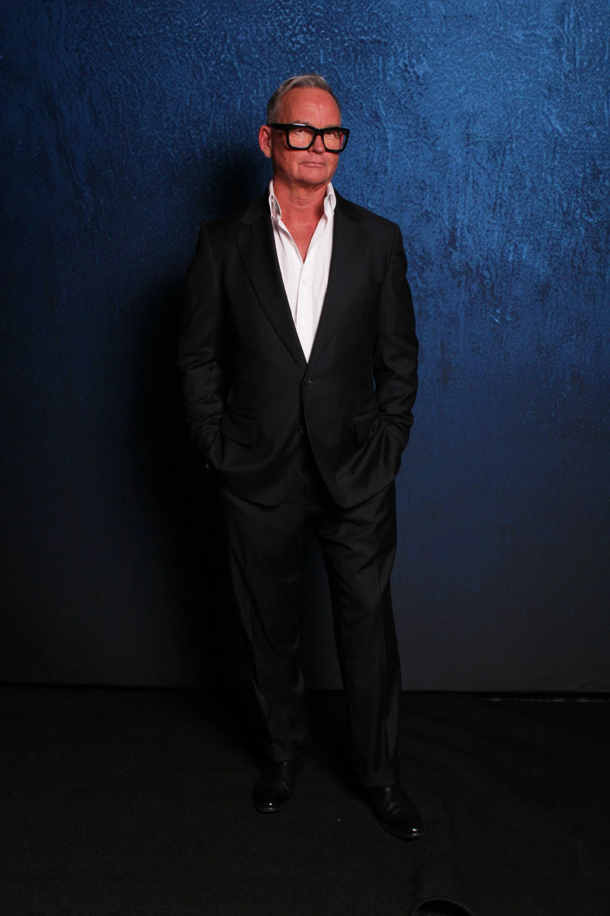 A man in a black suit, white shirt, and black shoes standing with hands in pockets against a dark textured blue background.