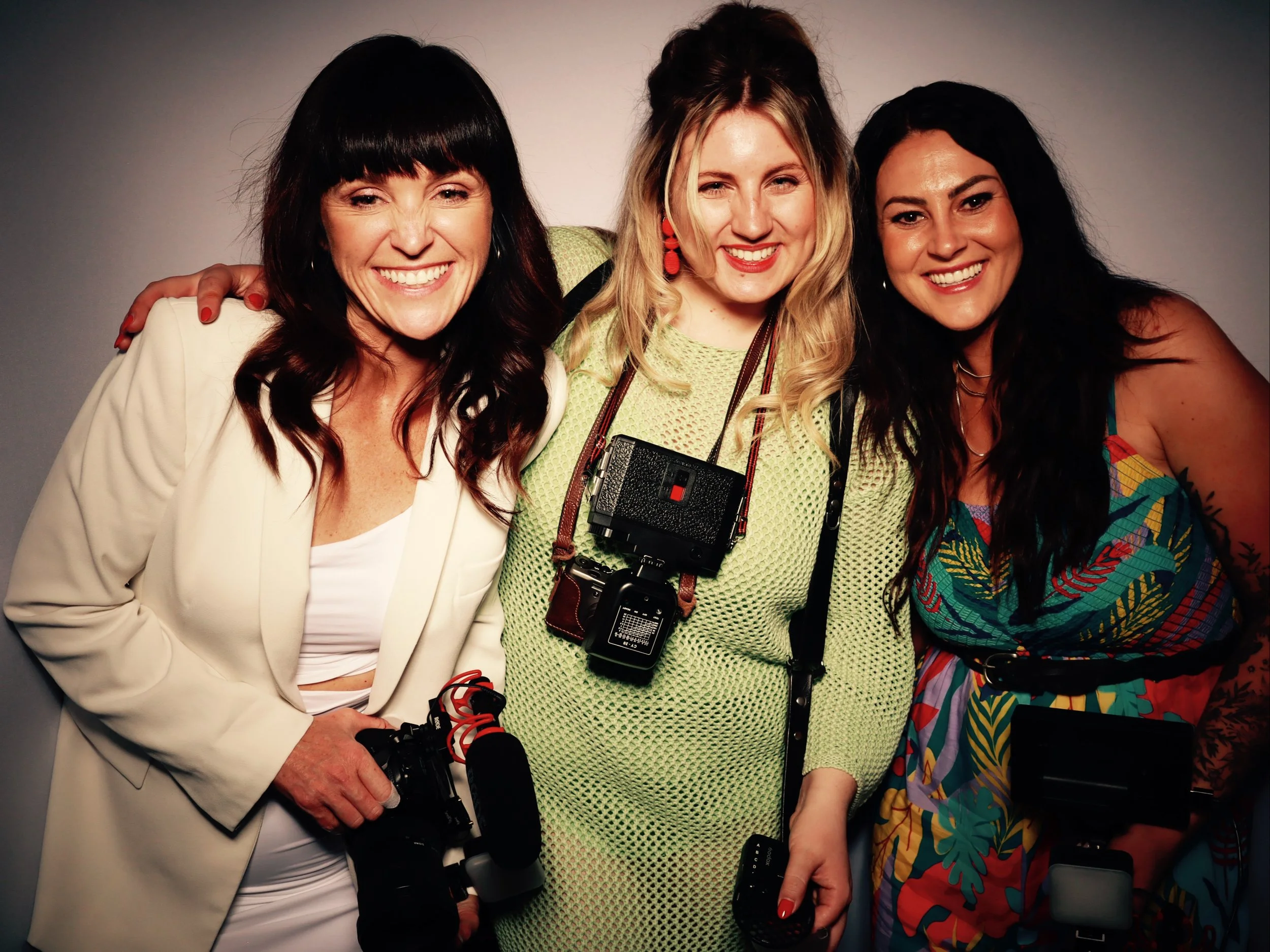 Three women smiling, standing close together, with cameras in their hands, on a plain background.