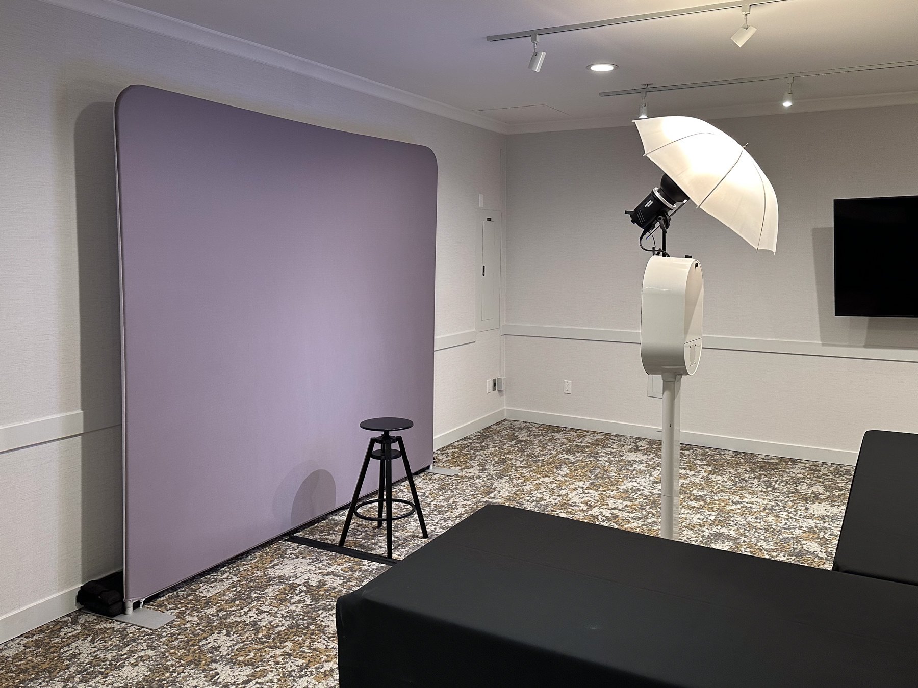 Interior of photography studio with large purple backdrop, photography light with umbrella, black stool, and a black table, tiled carpet, white walls, and a wall-mounted TV.