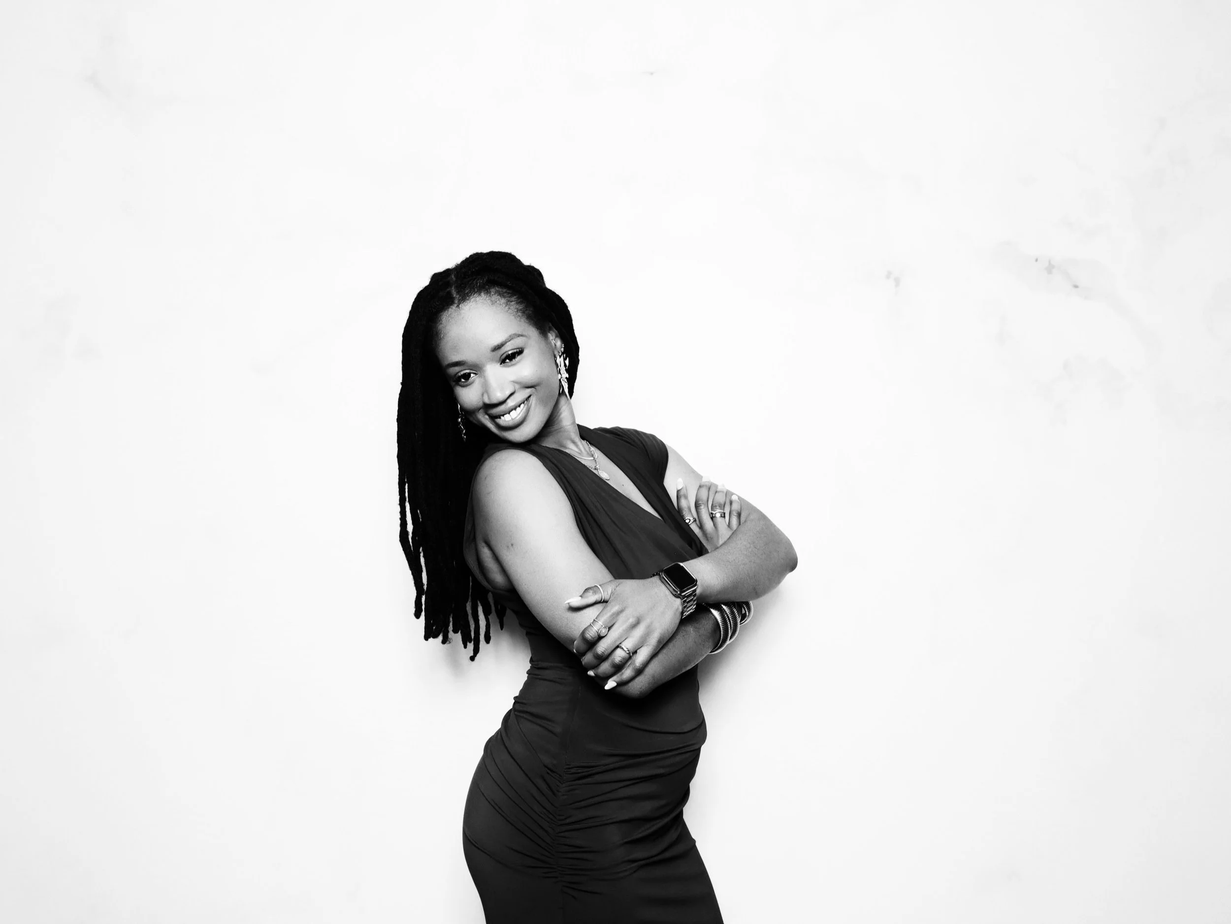 Black and white photo of a woman with long dreadlocks, wearing a sleeveless dark dress, smiling, with arms crossed, standing against a plain white background.
