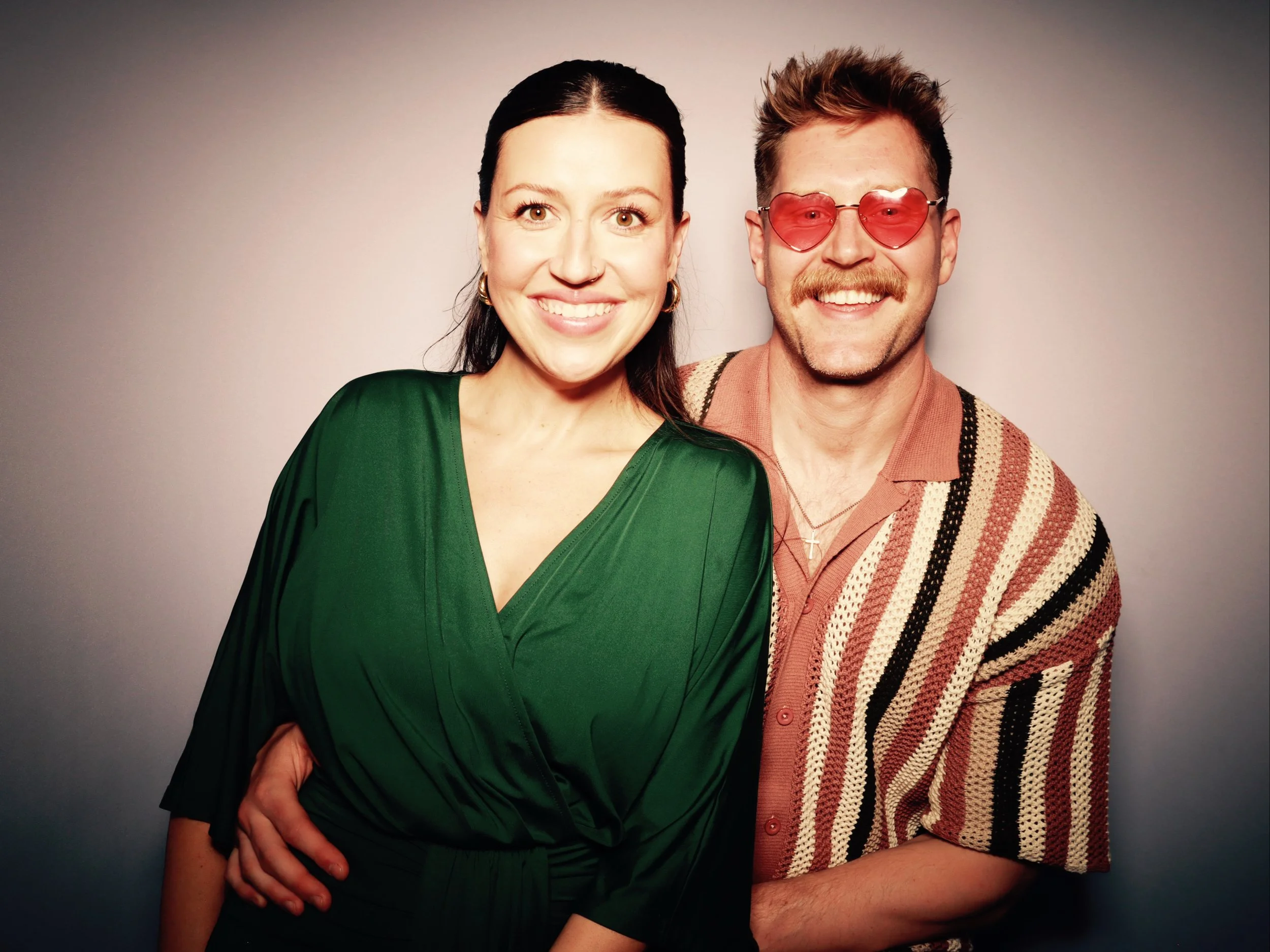A smiling woman in a green dress standing next to a man wearing heart-shaped sunglasses and a striped shirt, both posing against a plain background.