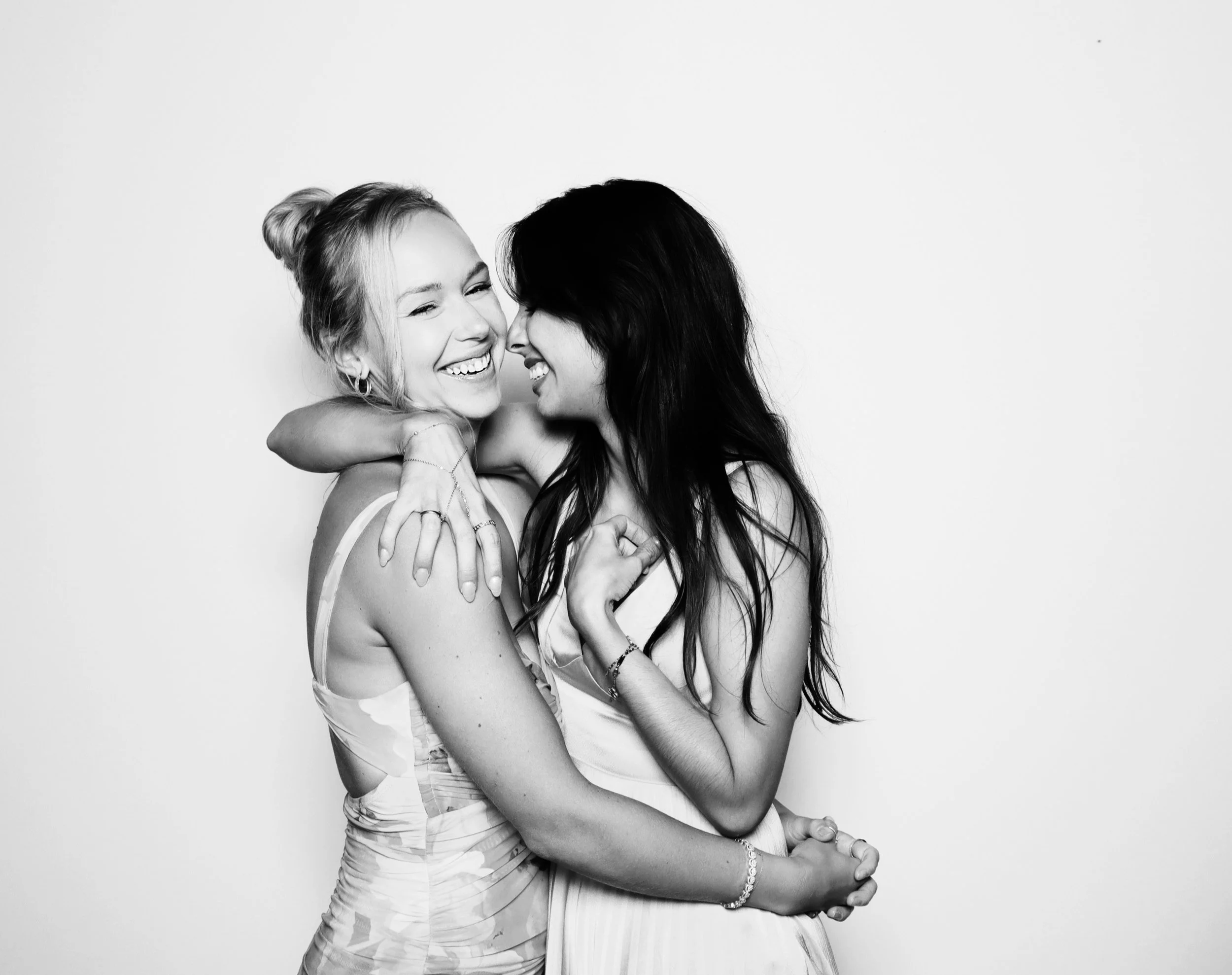 Two women are embracing and smiling, facing each other against a plain background.