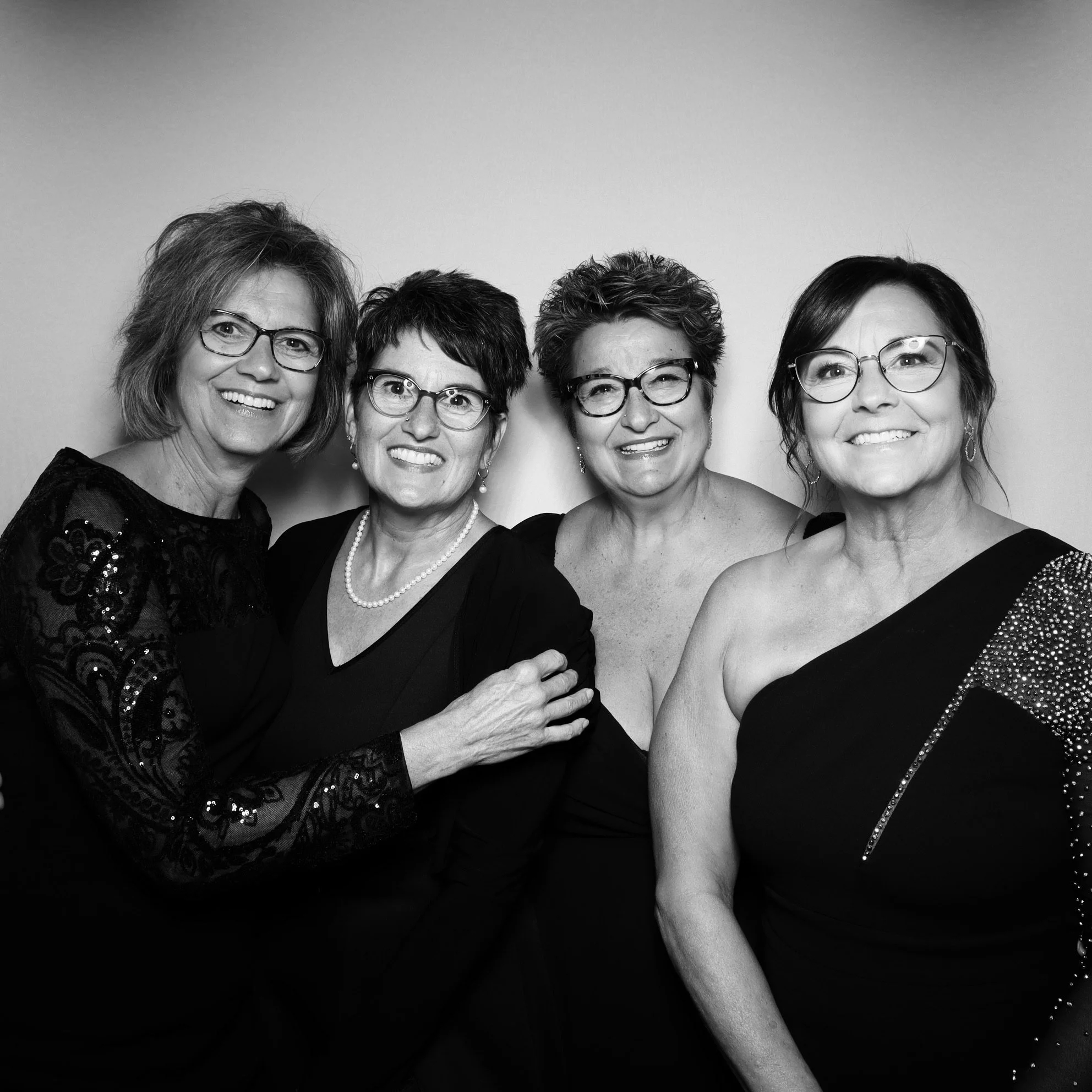 Black and white photo of four women dressed in elegant evening gowns and wearing glasses, smiling and posing together.
