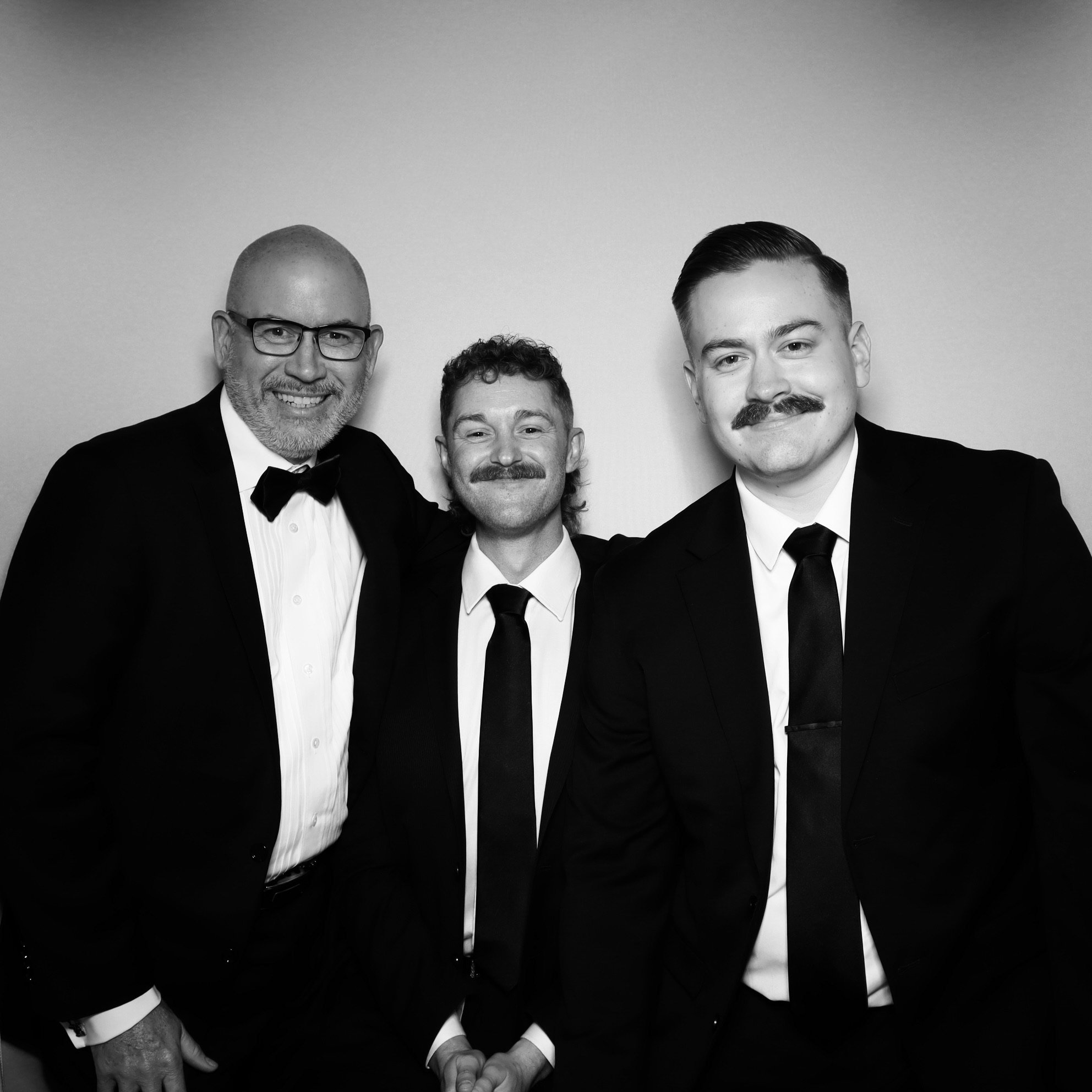 Three men in formal black tuxedos with ties, smiling, posing in front of a plain background in a black and white photograph.