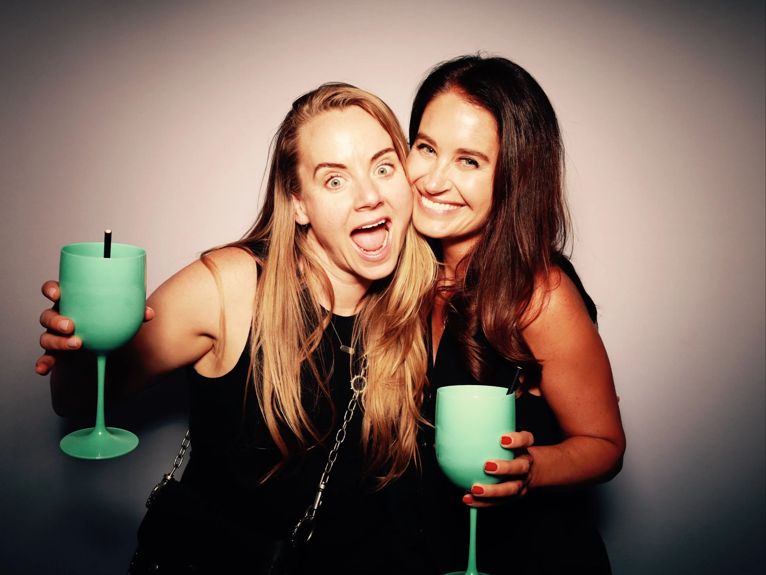 Two women smiling and posing together, each holding a large green drink with a straw.