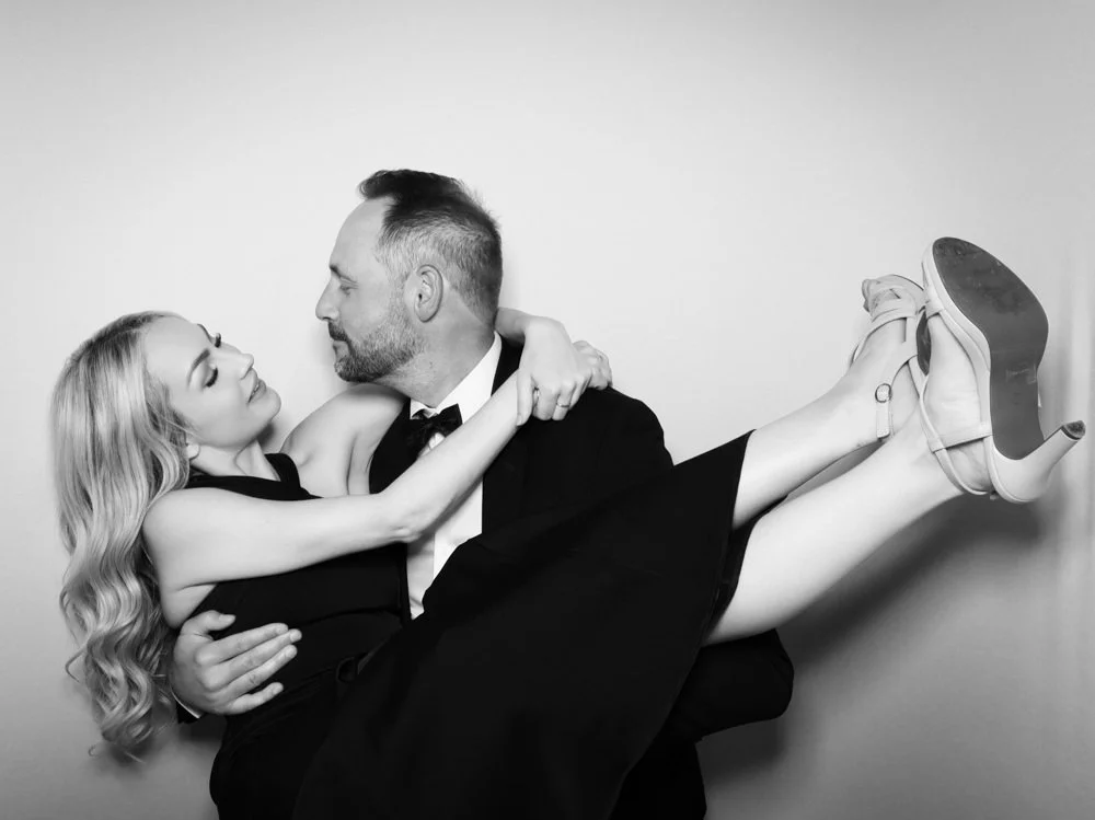 A black-and-white photo of a man in a tuxedo holding a woman in a dress and high heels, with the woman looking at the man as they pose together.