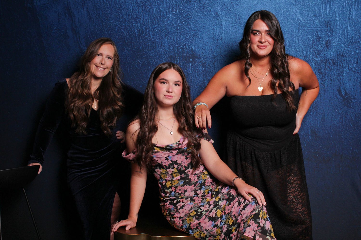 Three women with long, wavy brunette hair posing against a textured dark blue background. One woman is sitting on a chair in a floral dress, and two women are standing, one in a black dress and the other in a black strapless dress. All are smiling or
