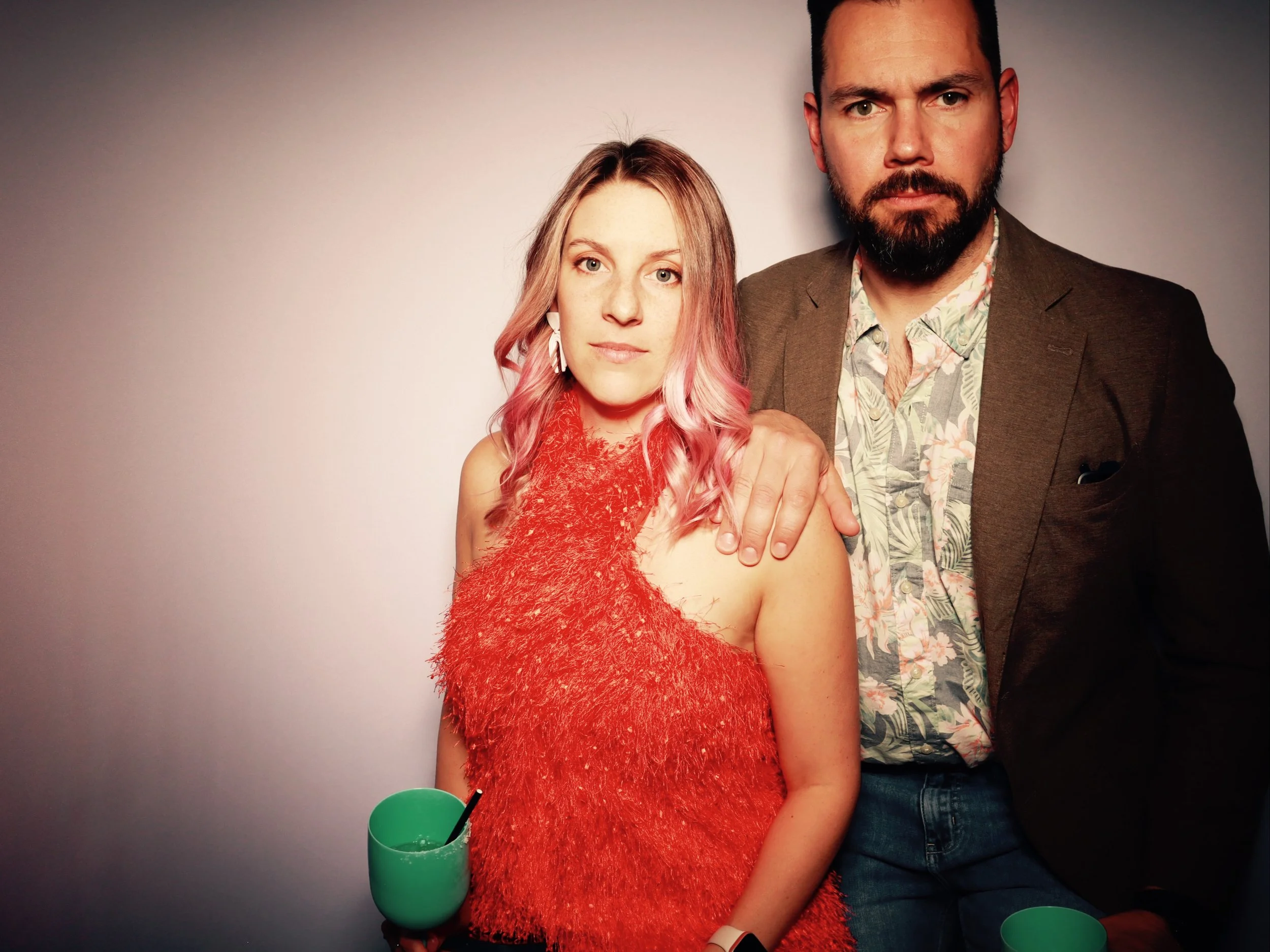 A young woman with pink hair and a red fuzzy dress stands next to a man with a beard, wearing a floral shirt and brown jacket. They are against a plain background, and the woman is holding a green cup with a straw.