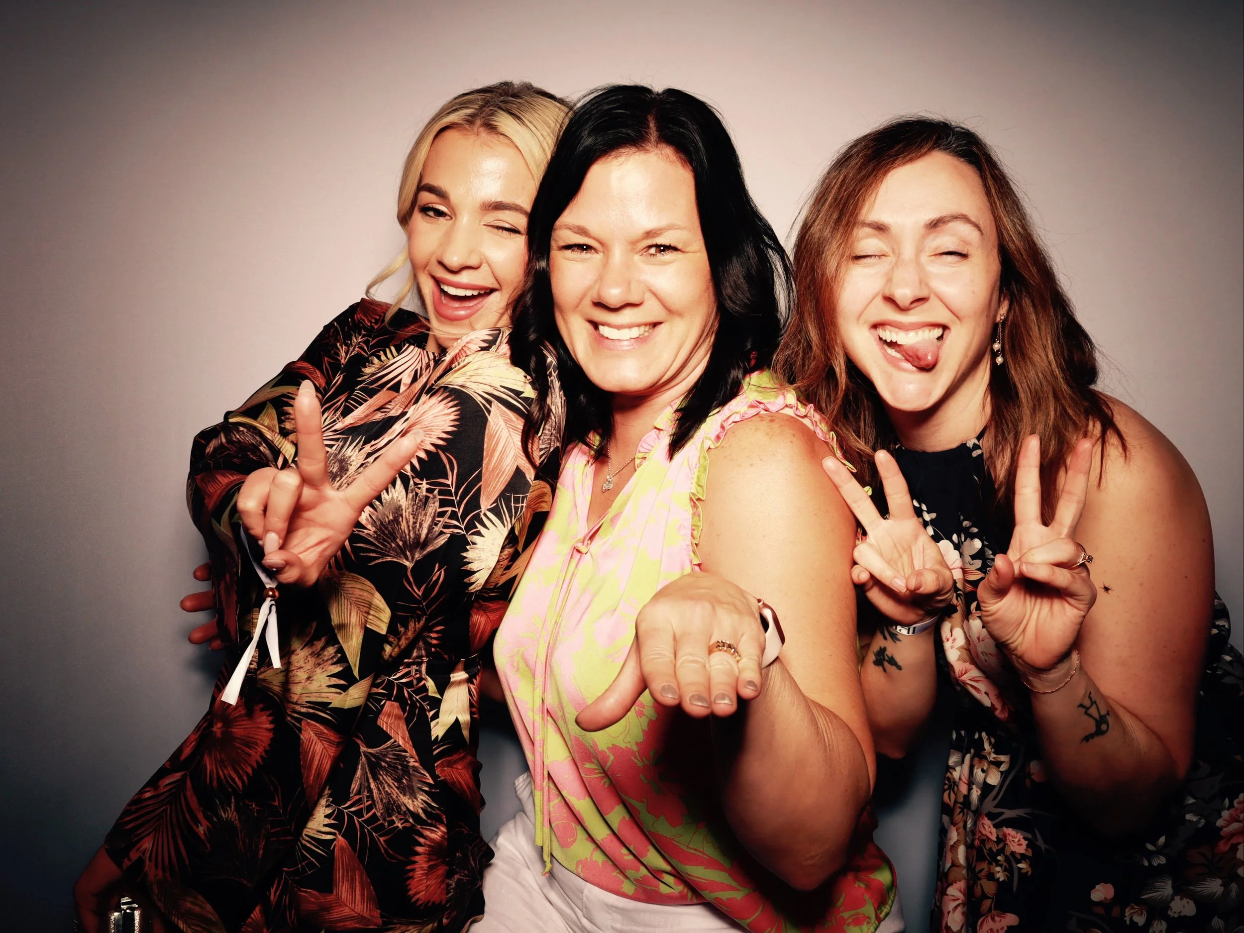 Three women posing together, making peace signs and playful faces, against a plain background.