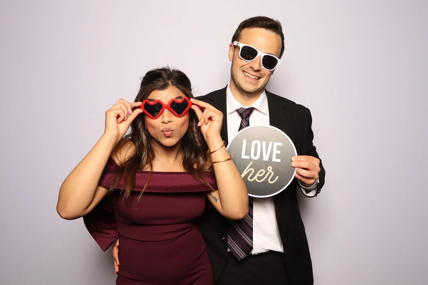 A man and woman posing together at a photo booth, with the woman wearing heart-shaped sunglasses and the man wearing sunglasses, holding a sign that says 'Love her'.