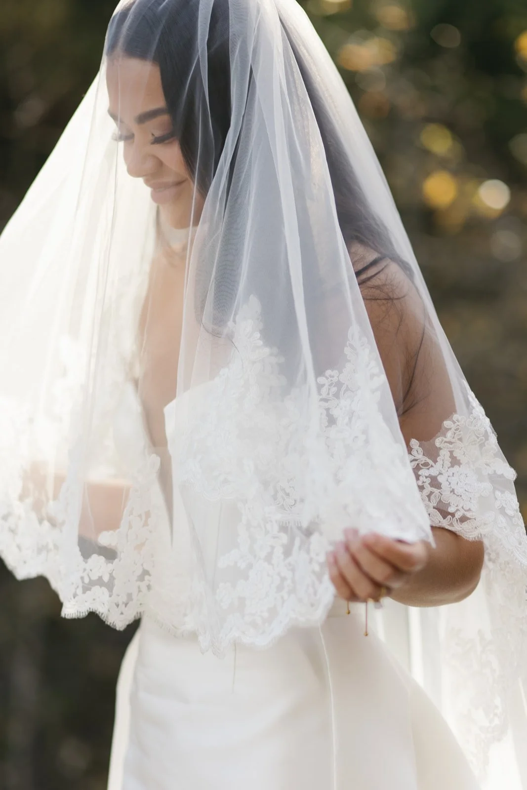 A bride with dark hair in a white wedding dress, wearing a lace veil, smiling and looking down, outdoors with blurred trees in the background.