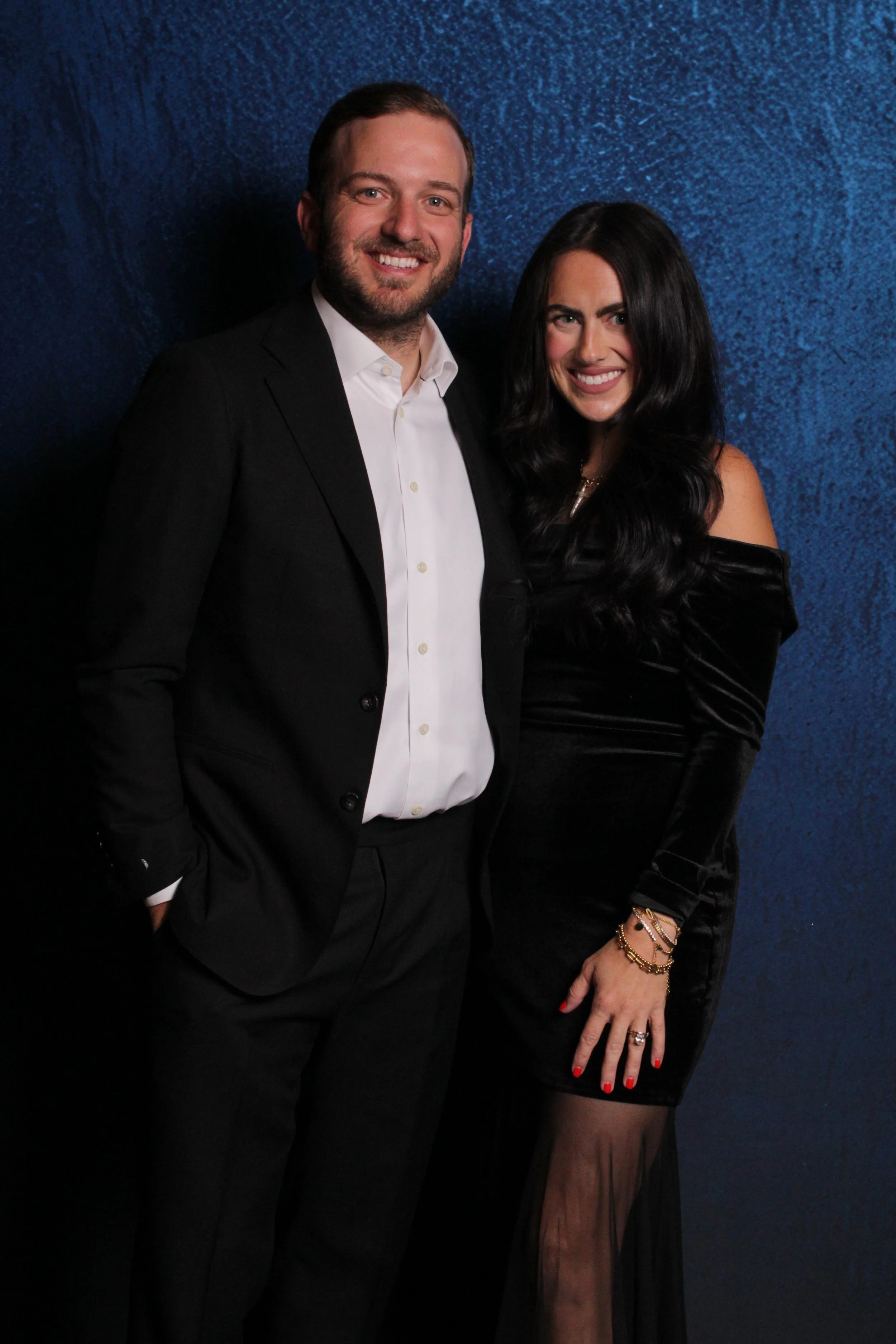 A man in a black suit and white shirt stands next to a woman in a black off-shoulder dress with long black hair, posing against a textured blue background.