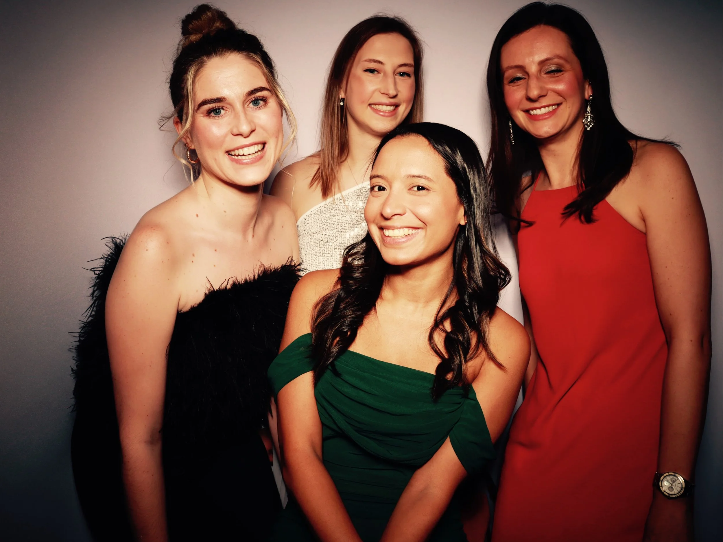 Group of five women dressed in elegant evening attire standing close together and smiling at the camera, against a plain background.