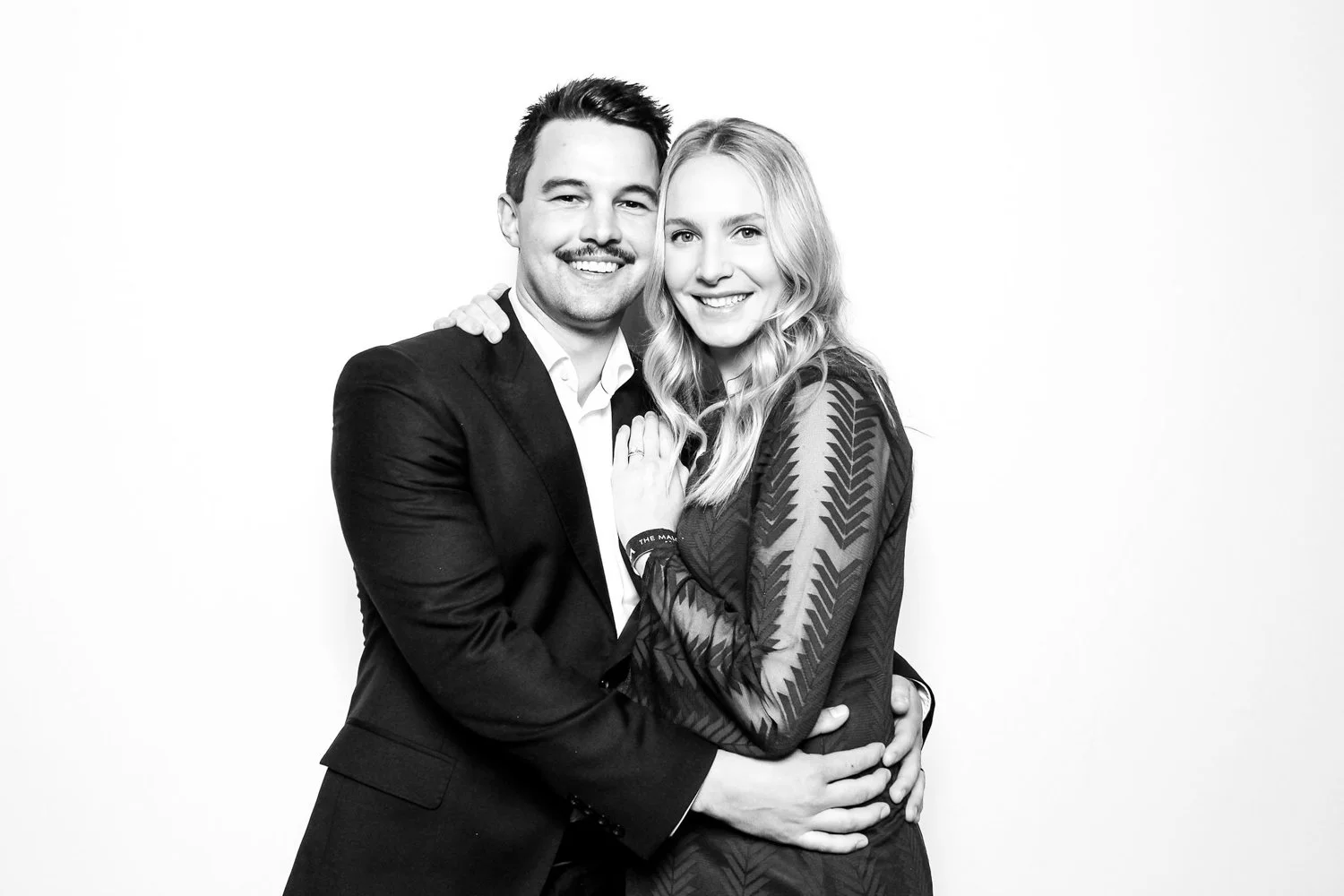 A smiling man and woman hugging each other against a plain white background. The man has short dark hair and a mustache, wearing a dark suit with a white shirt. The woman has long wavy blonde hair and is wearing a patterned long-sleeve top.