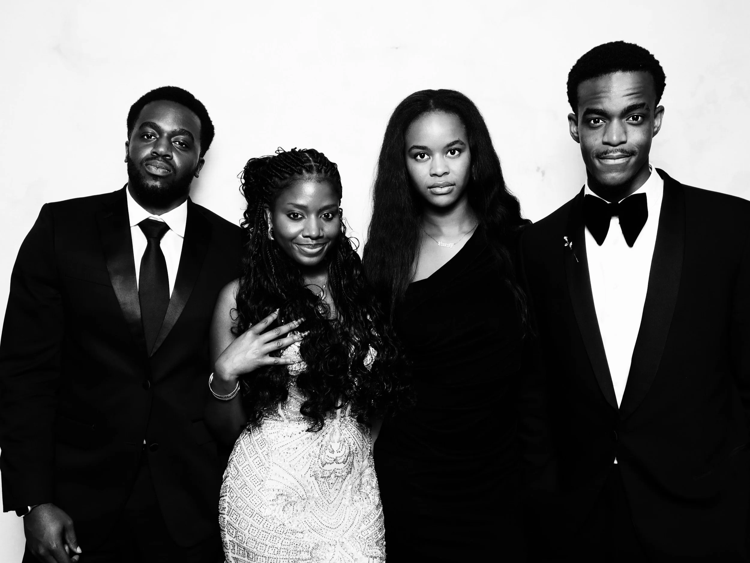 Four people dressed in formal attire standing together against a plain background, two men in tuxedos and two women in elegant dresses.