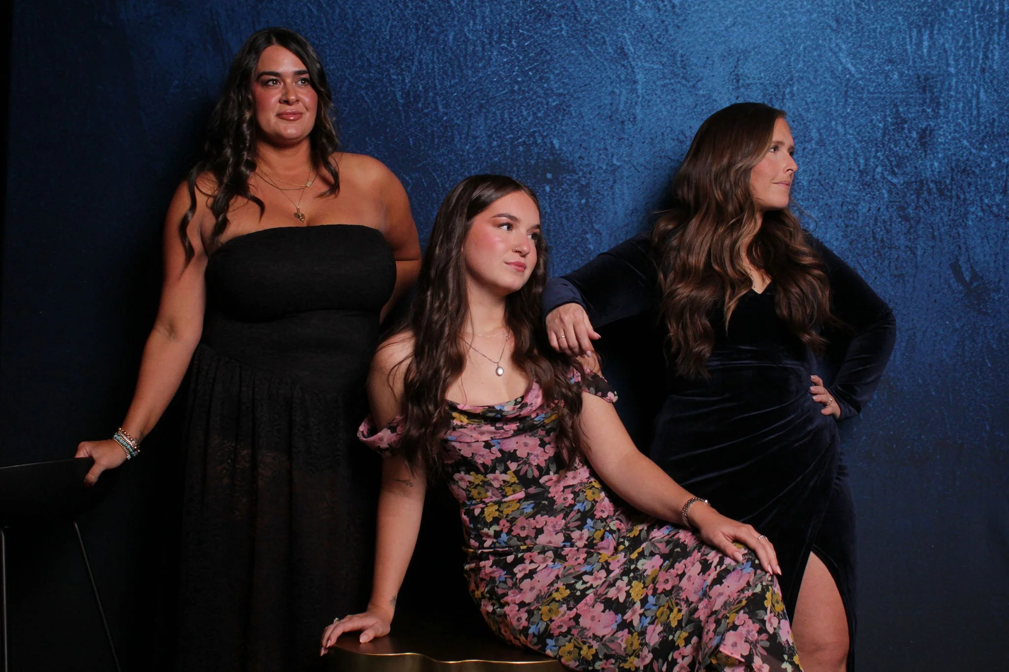 Three women posing in front of a dark blue wall, two standing and one sitting on a black chair, wearing elegant dresses.