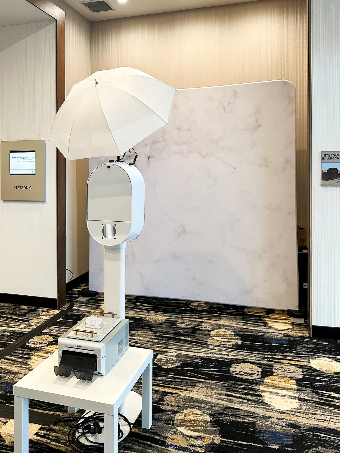 Photo of a photo booth with a white umbrella, a machine on a white table, and a reflective backdrop in a hotel or conference center hallway.