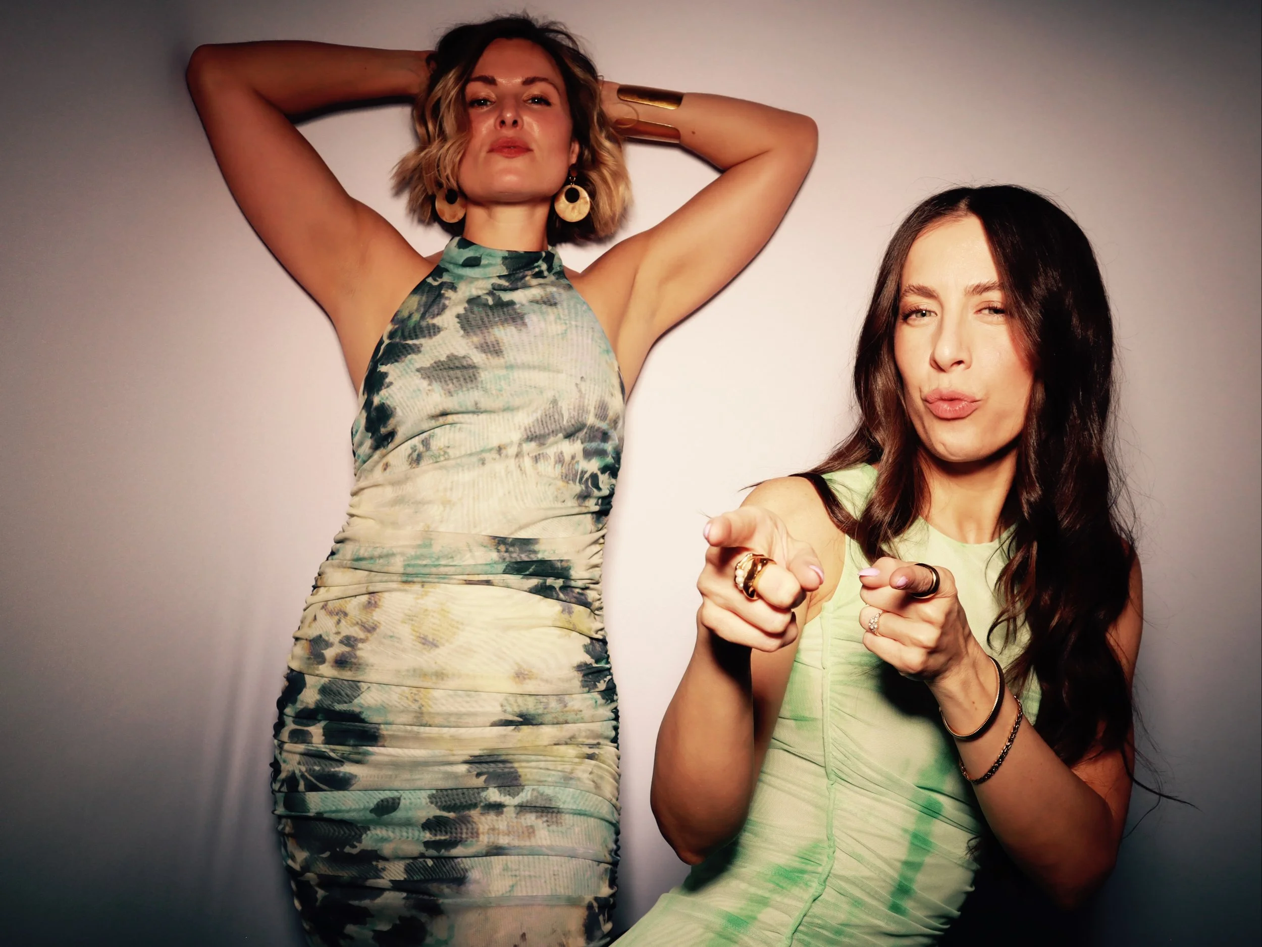 Two women posing against a plain wall, one with hands behind her head wearing a patterned dress, the other with a playful expression pointing at the camera wearing a light green dress.