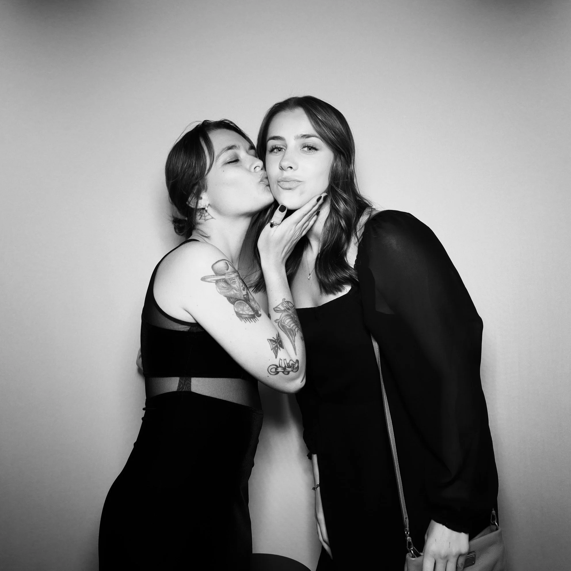 Two women, one kissing the other's cheek, the second woman is smiling, both dressed in dark clothing, standing against a plain background.