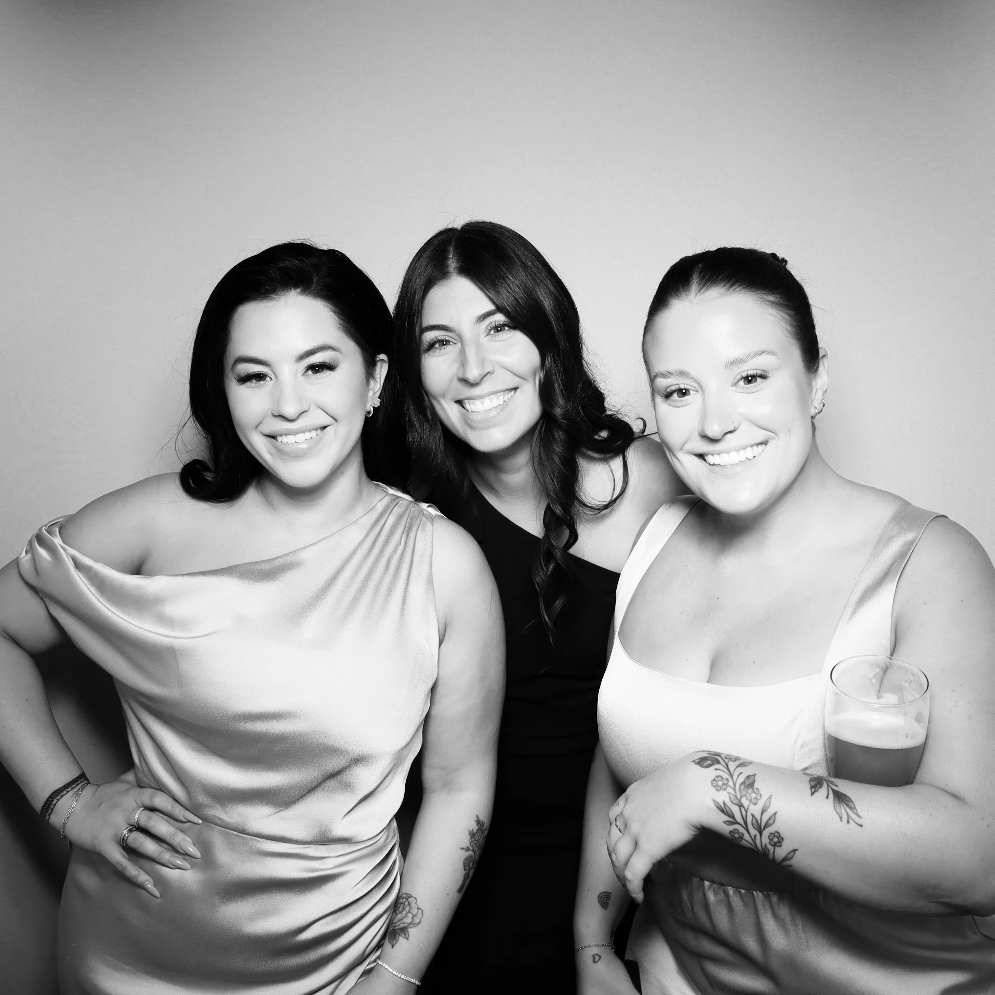 Three women smiling and posing together, one holding a drink, in black and white photo.