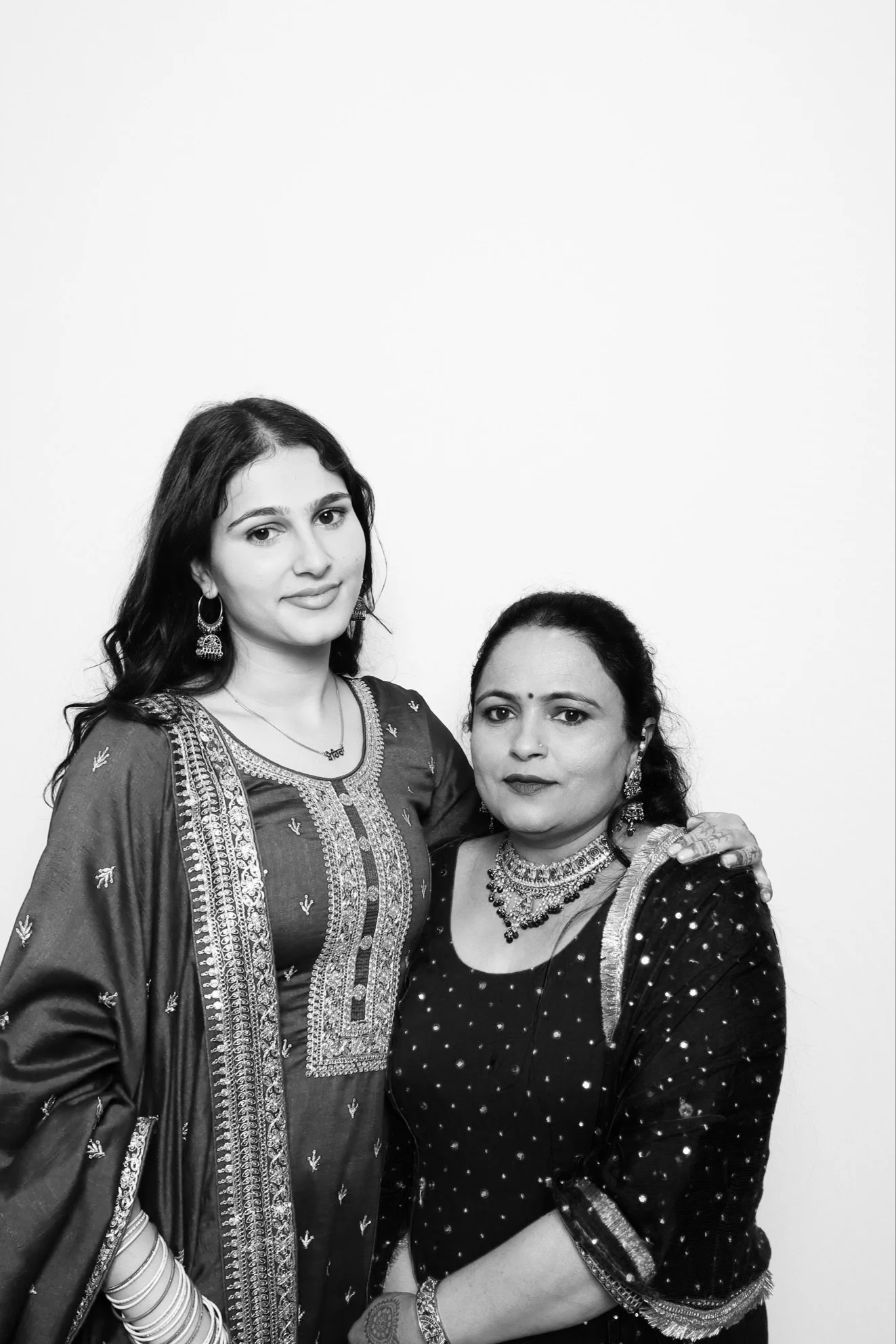 Black and white photo of two women wearing traditional Indian clothing and jewelry, standing close together against a plain white background.