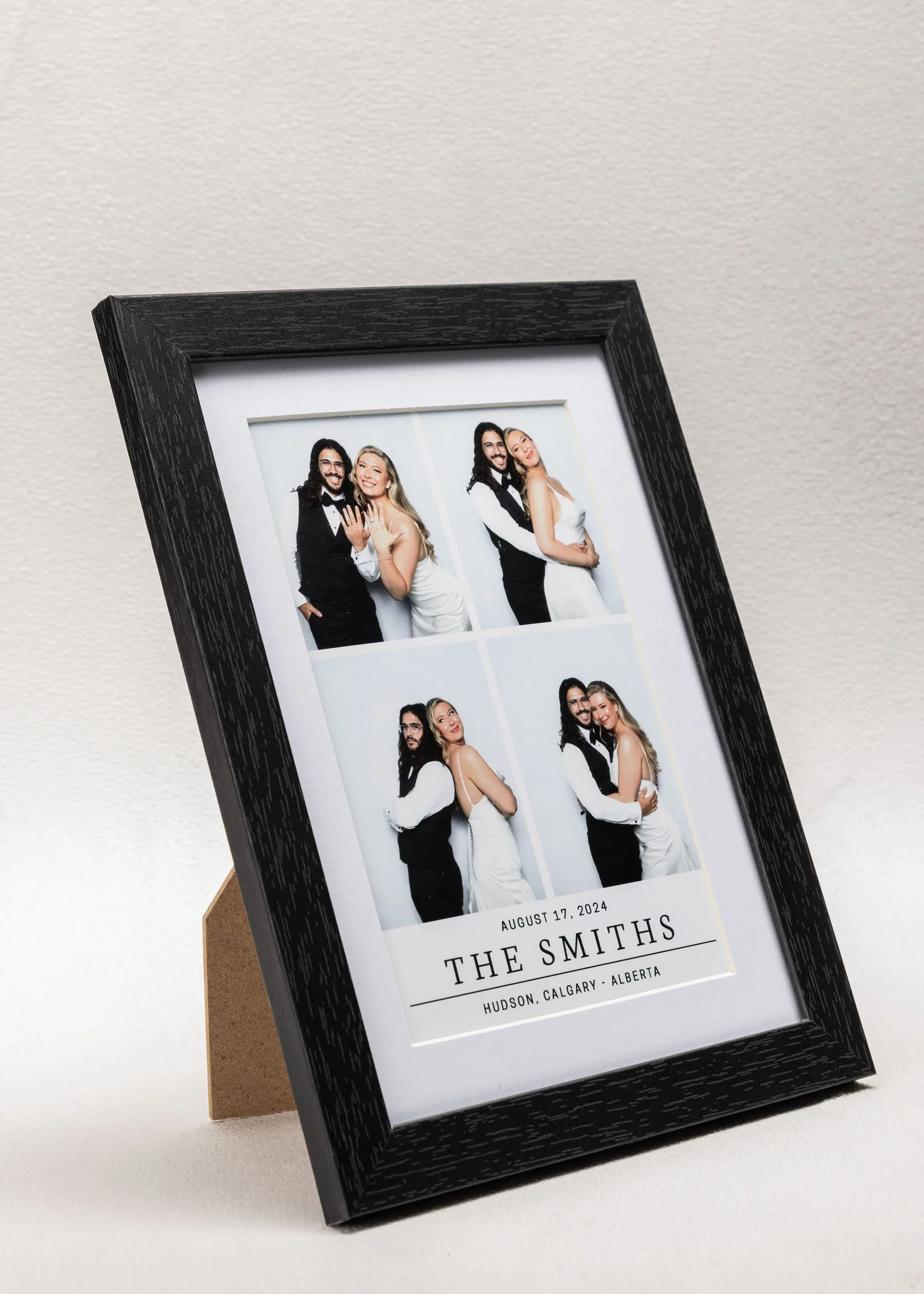 Photo booth strip with four pictures of a couple, date August 17, 2024, and text 'The Smiths, Hudson, Calgary, Alberta', displayed in a black frame on a beige surface.