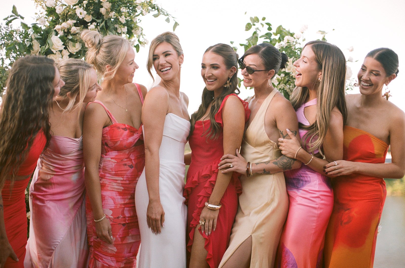 A group of nine women dressed in colorful dresses, smiling, and standing close together outdoors at a celebration.