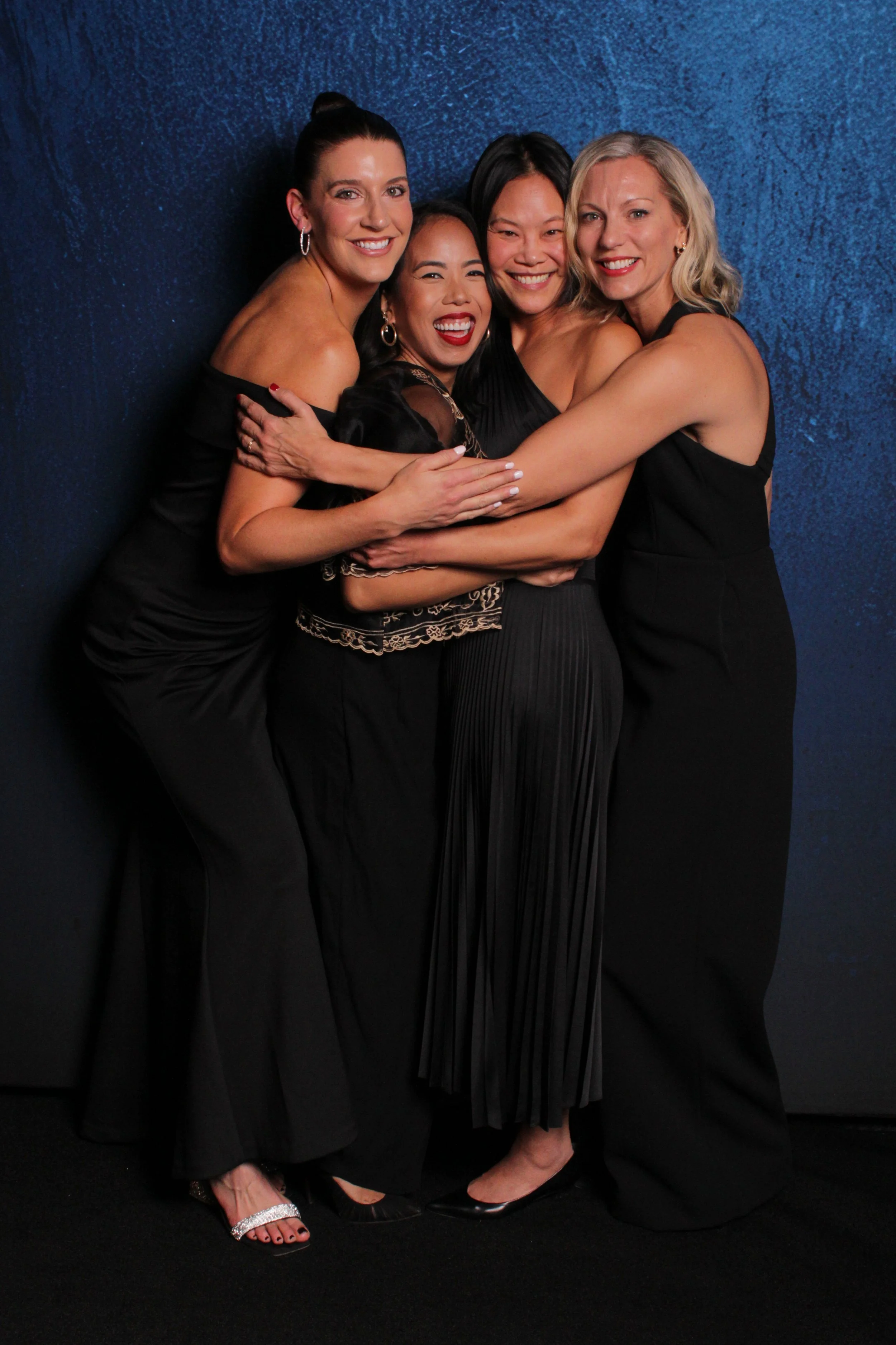 Four women hugging together, smiling, dressed in black evening wear, standing against a blue textured wall.