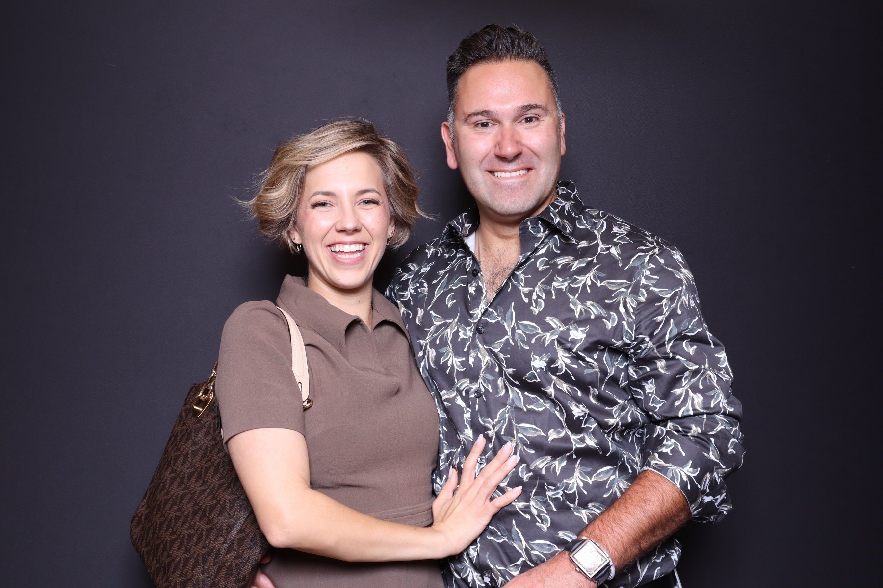 A smiling couple posing together against a black background. The woman has short blond hair, wearing a brown blouse and carrying a patterned purse. The man has short dark hair, wearing a black shirt with a white floral pattern, and a watch.