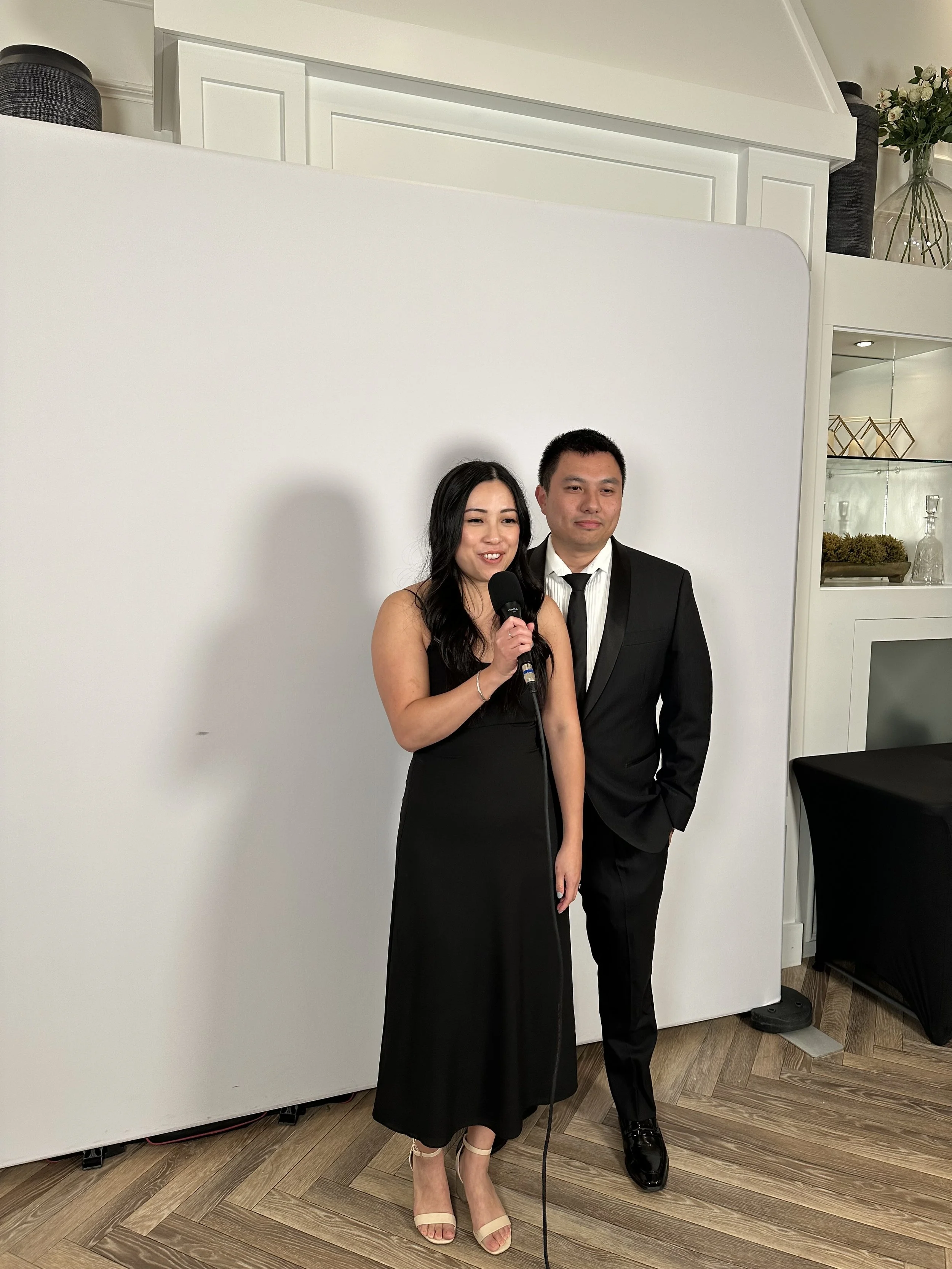 A woman in a black dress holding a microphone stands next to a man in a black suit at an indoor event with a plain white backdrop and decorative decor in the background.