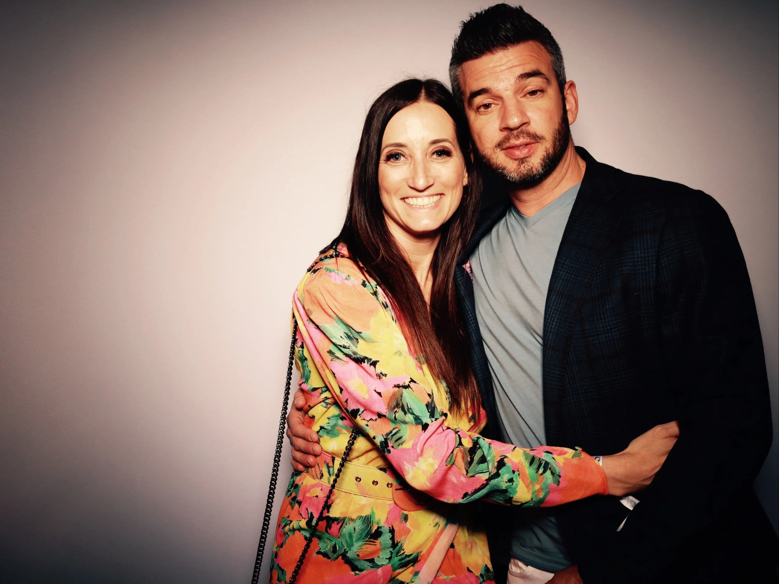 A smiling woman with long dark hair and a man with short dark hair and beard, embracing each other against a plain wall. The woman is wearing a colorful floral dress with pink, yellow, and green patterns, and the man is dressed in a black jacket over